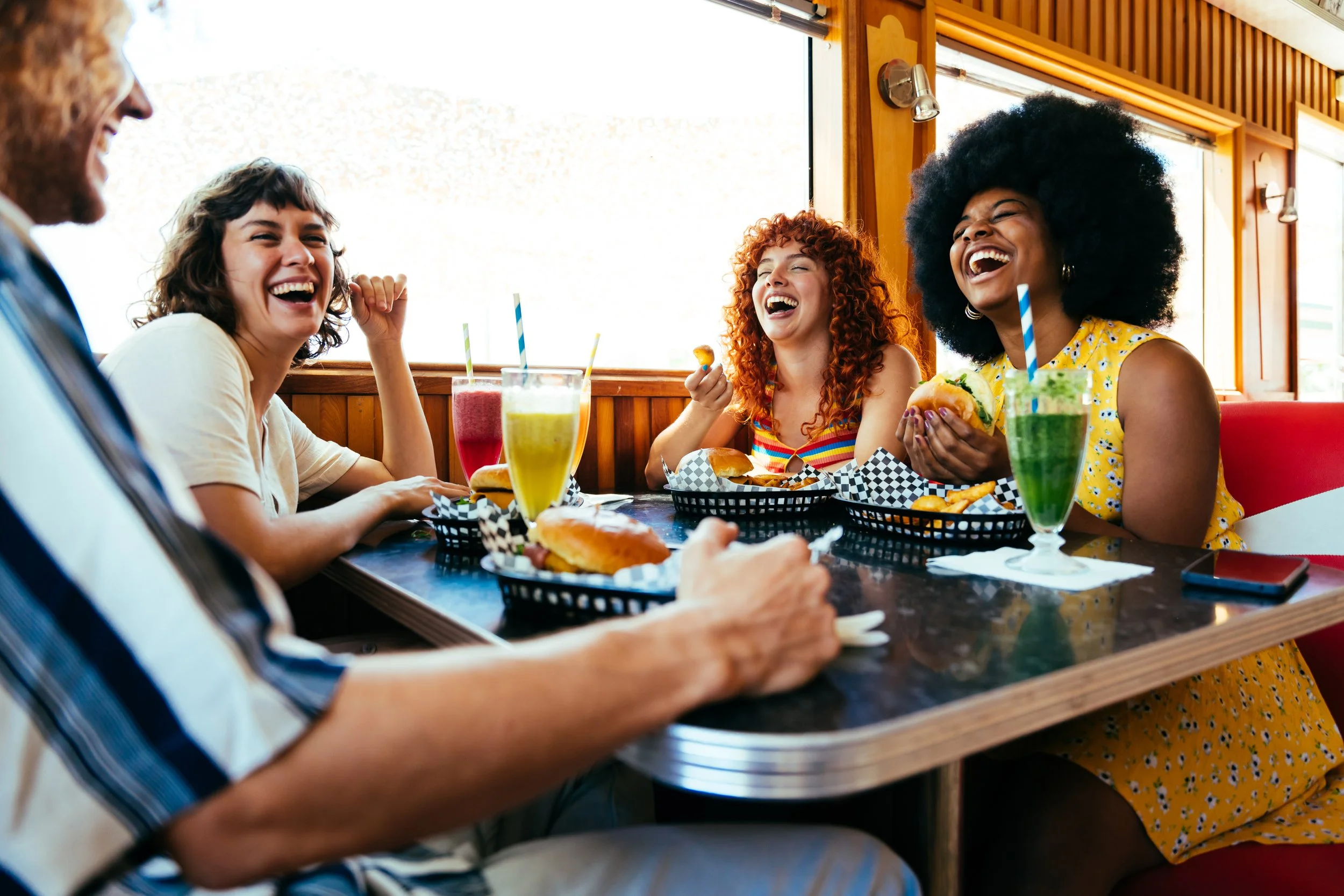 Four women are sitting at a table in a restaurant, laughing and enjoying themselves. They have hamburgers and colorful drinks with straws in front of them, and there's a person in a car seat visible in the foreground.