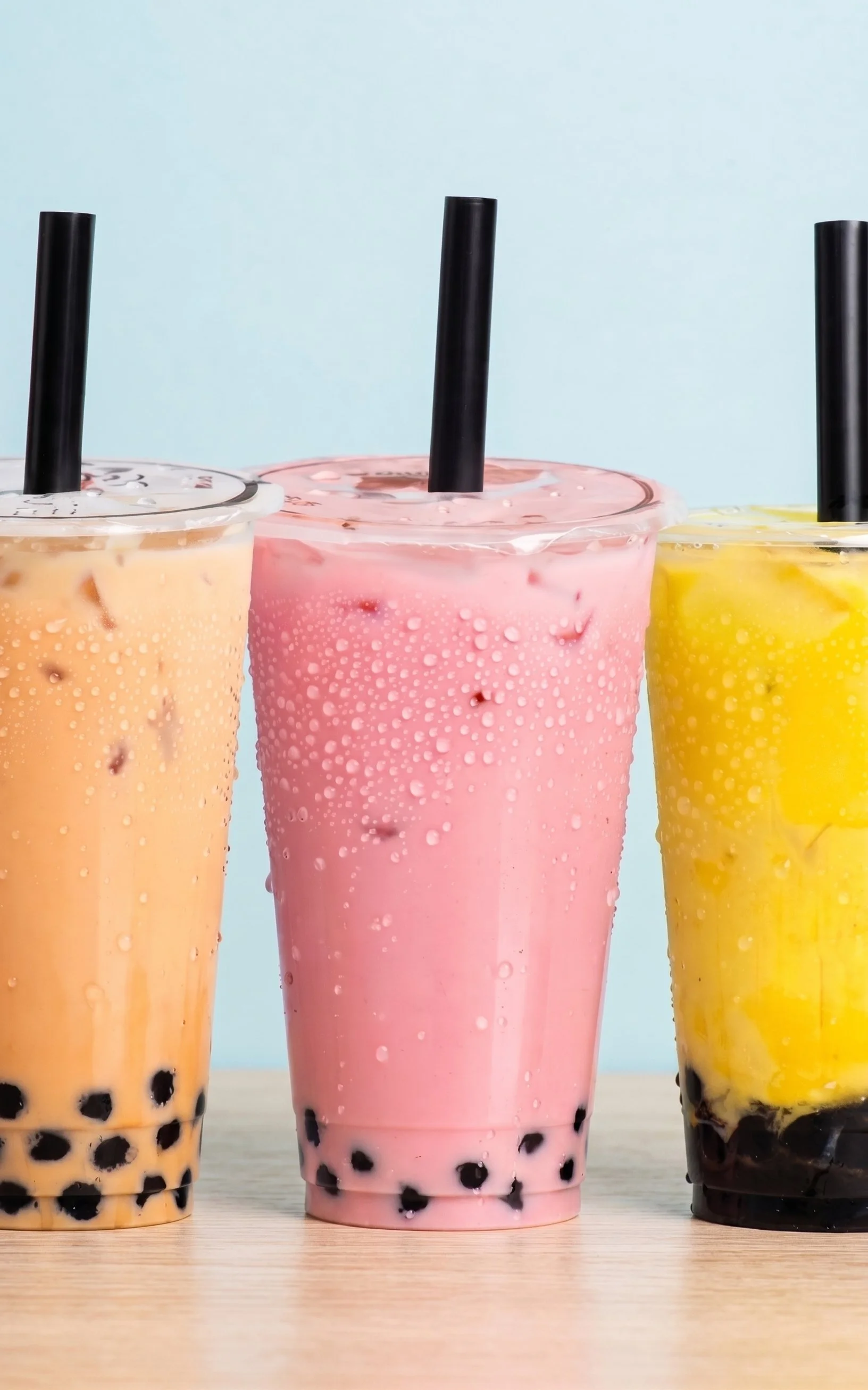 Three large cups of bubble tea with different flavors, one peach, one pink, and one yellow, with black tapioca pearls at the bottom and black straws on a wooden surface against a light blue background.
