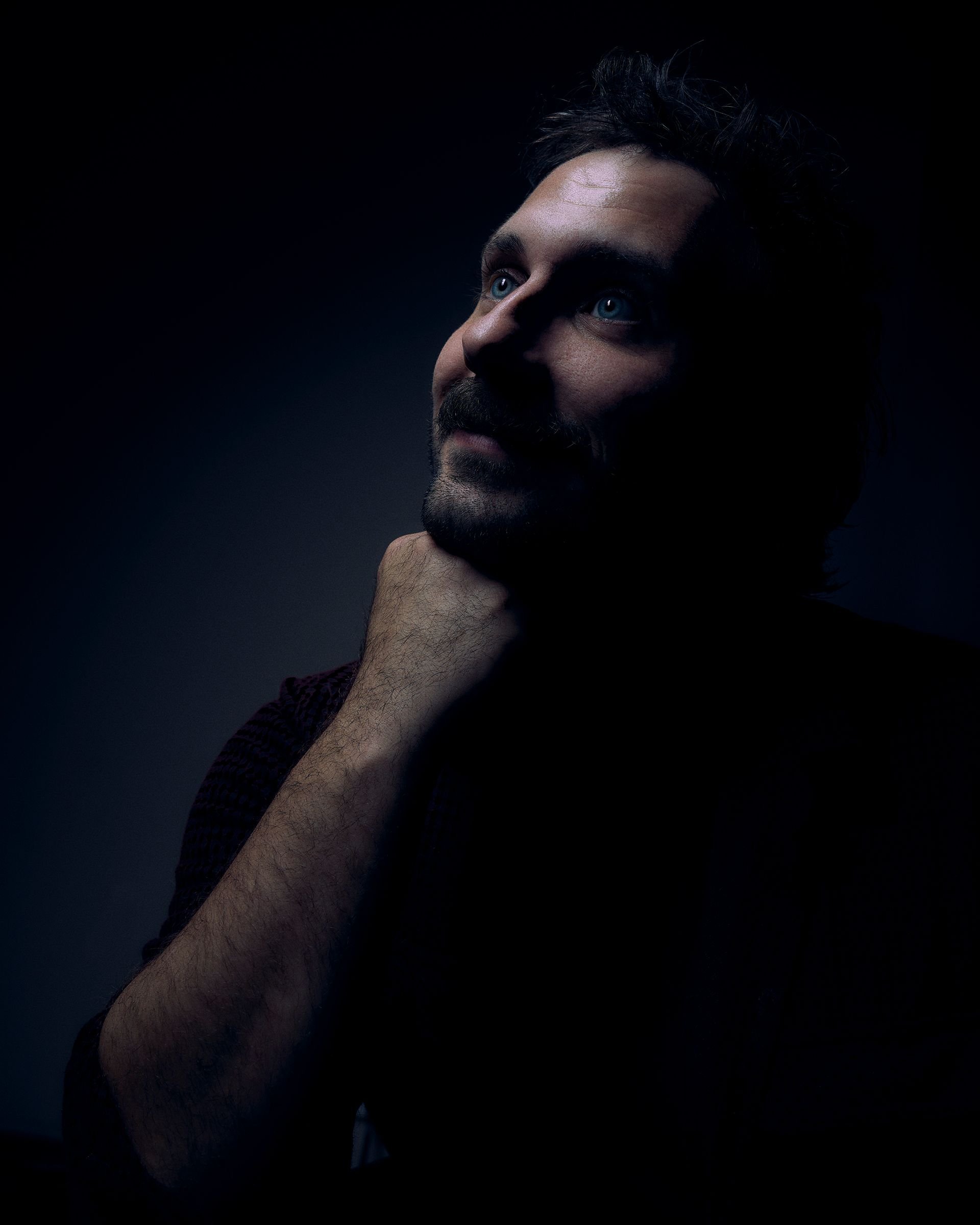A man with dark hair and a beard, looking upward, is partially illuminated against a dark background. His hand rests on his neck.