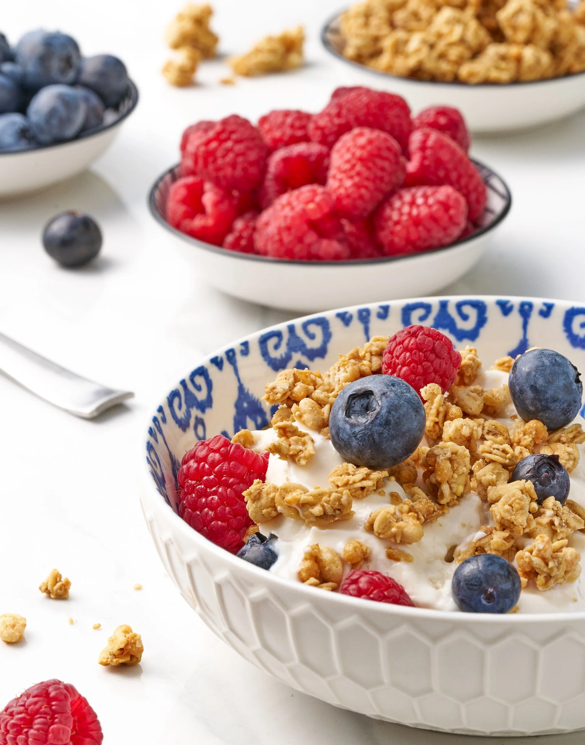 A close-up of a bowl of yogurt topped with granola, blueberries, and raspberries, with similar bowls and scattered granola in the background.