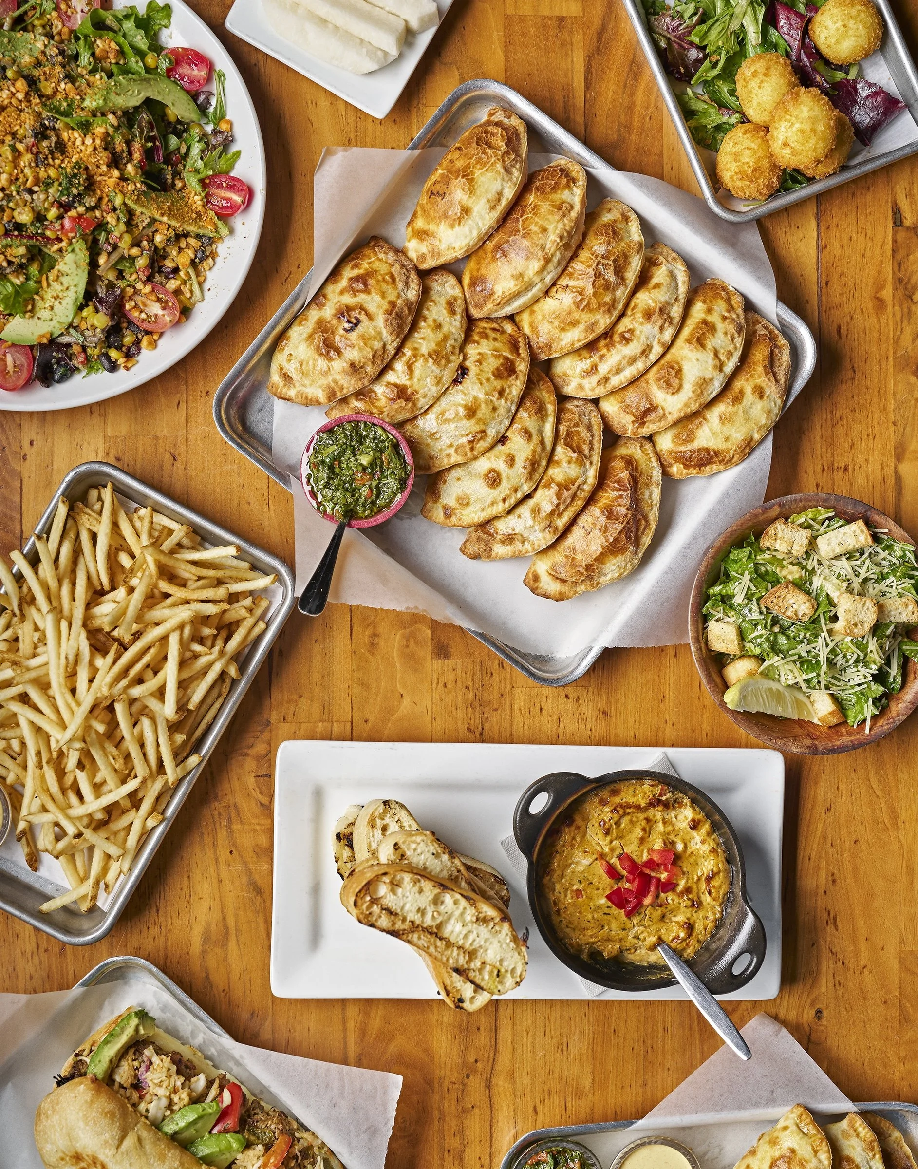 A variety of dishes on a wooden table including empanadas, a salad, French fries, and a bowl of baked cheese dip with red bell peppers, along with other small plates of food.