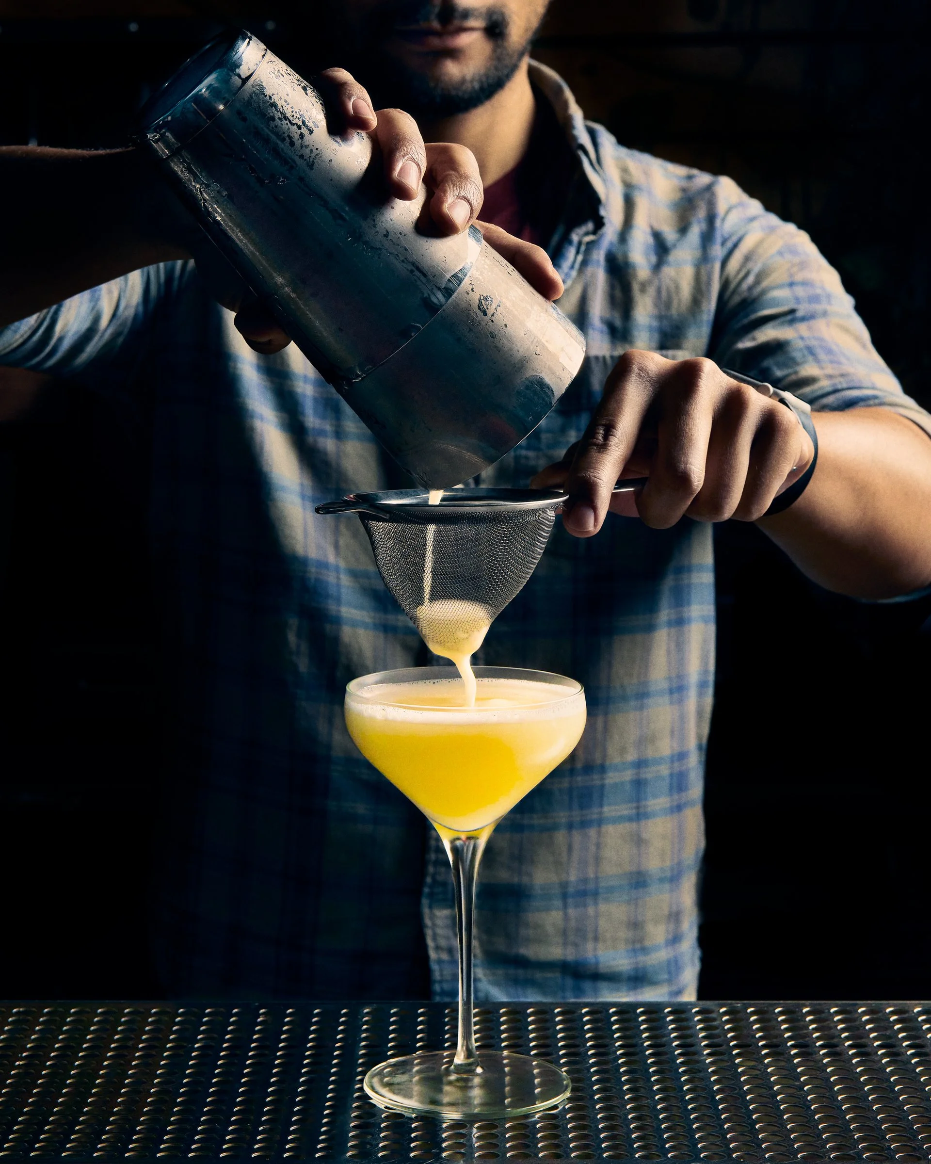A man in a checkered shirt is pouring a yellow cocktail from a shaker through a strainer into a cocktail glass. The scene appears to be in a bar with a textured black surface.