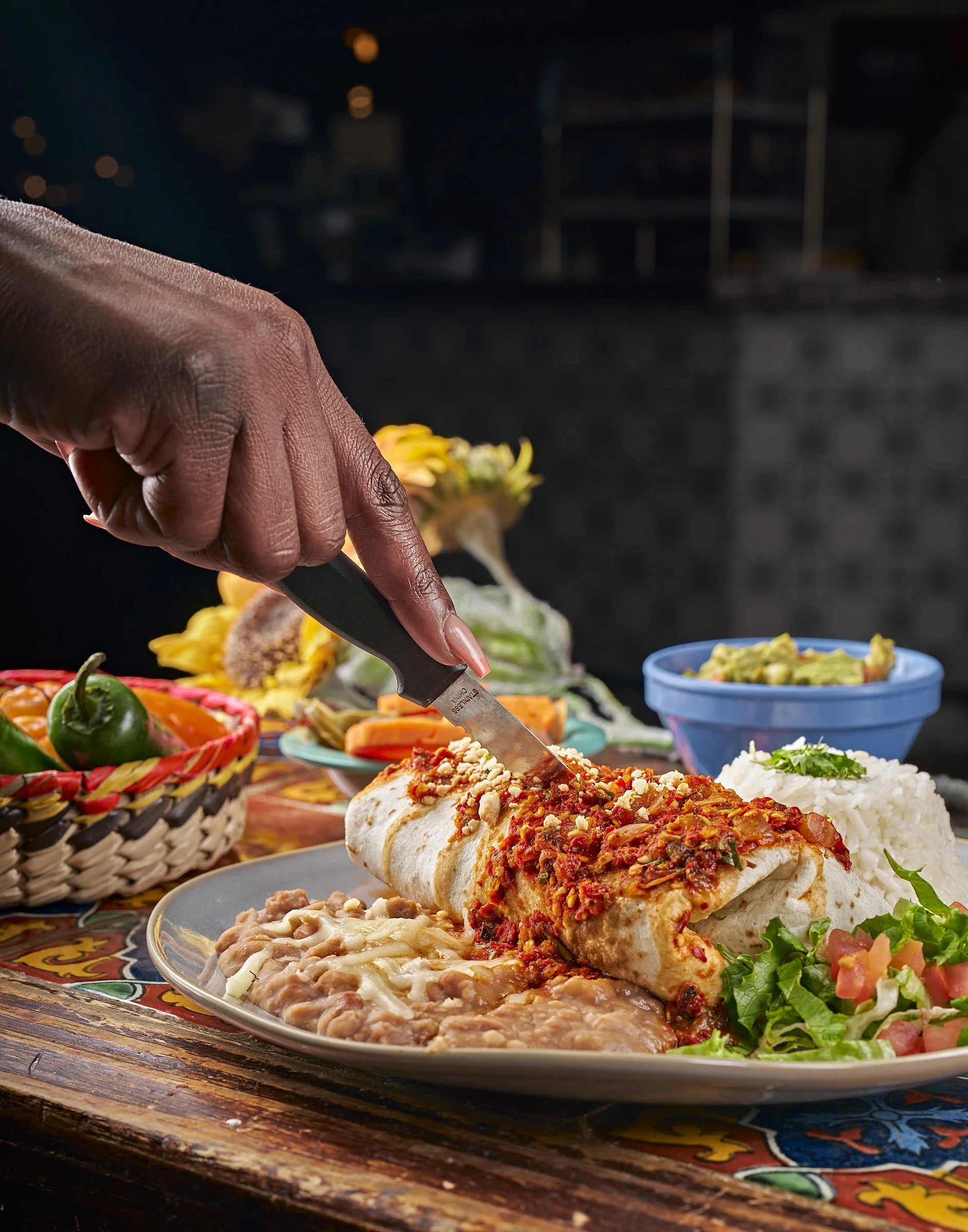 Mexican food plate with a burrito topped with red chili sauce and cheese, white rice, beans, and side salads surrounded by traditional colorful Mexican patterns.
