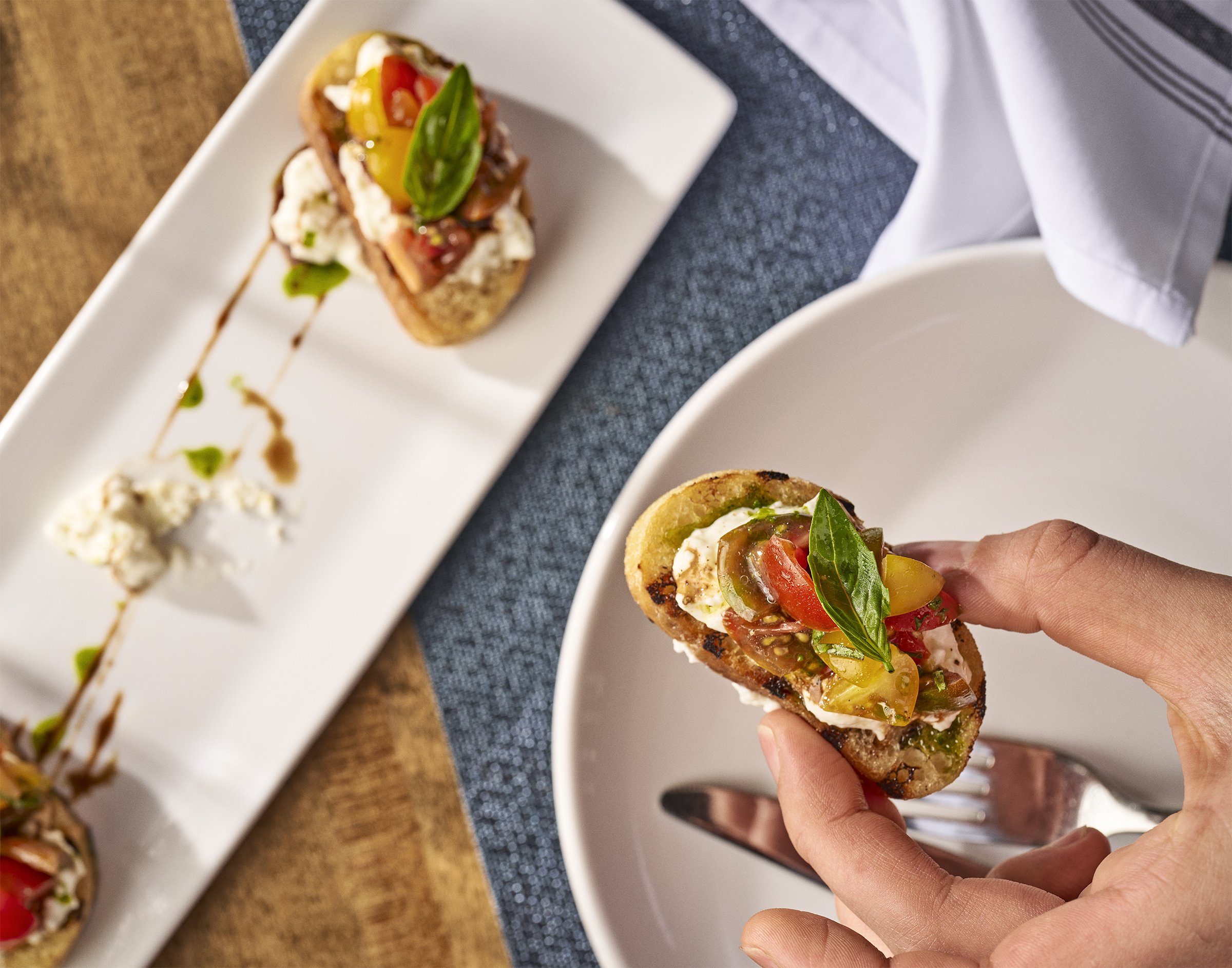 Person holding a slice of bruschetta topped with chopped tomatoes, basil, and mozzarella cheese on a dotted blue napkin, with more bruschetta on a white rectangular plate in the background.