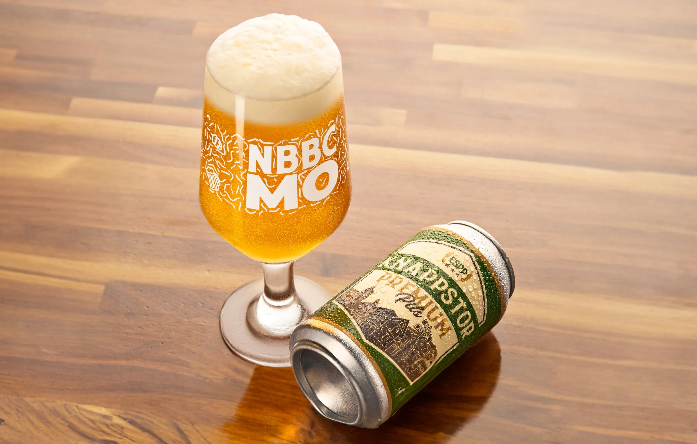 A beer glass with foam and the text 'NBC MO' on it, next to a can of New Barons Brewing Coop  beer lying on a wooden surface.