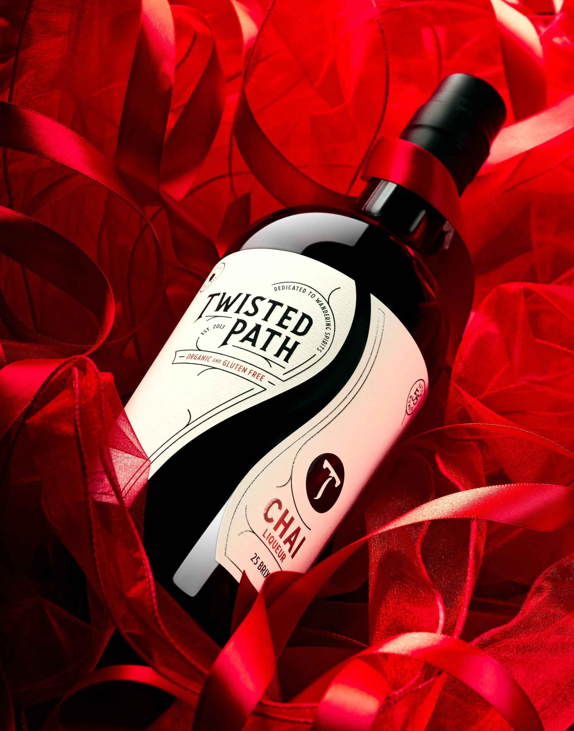 A wine bottle labeled 'Twisted Path' resting among red satin ribbons and red tissue paper.