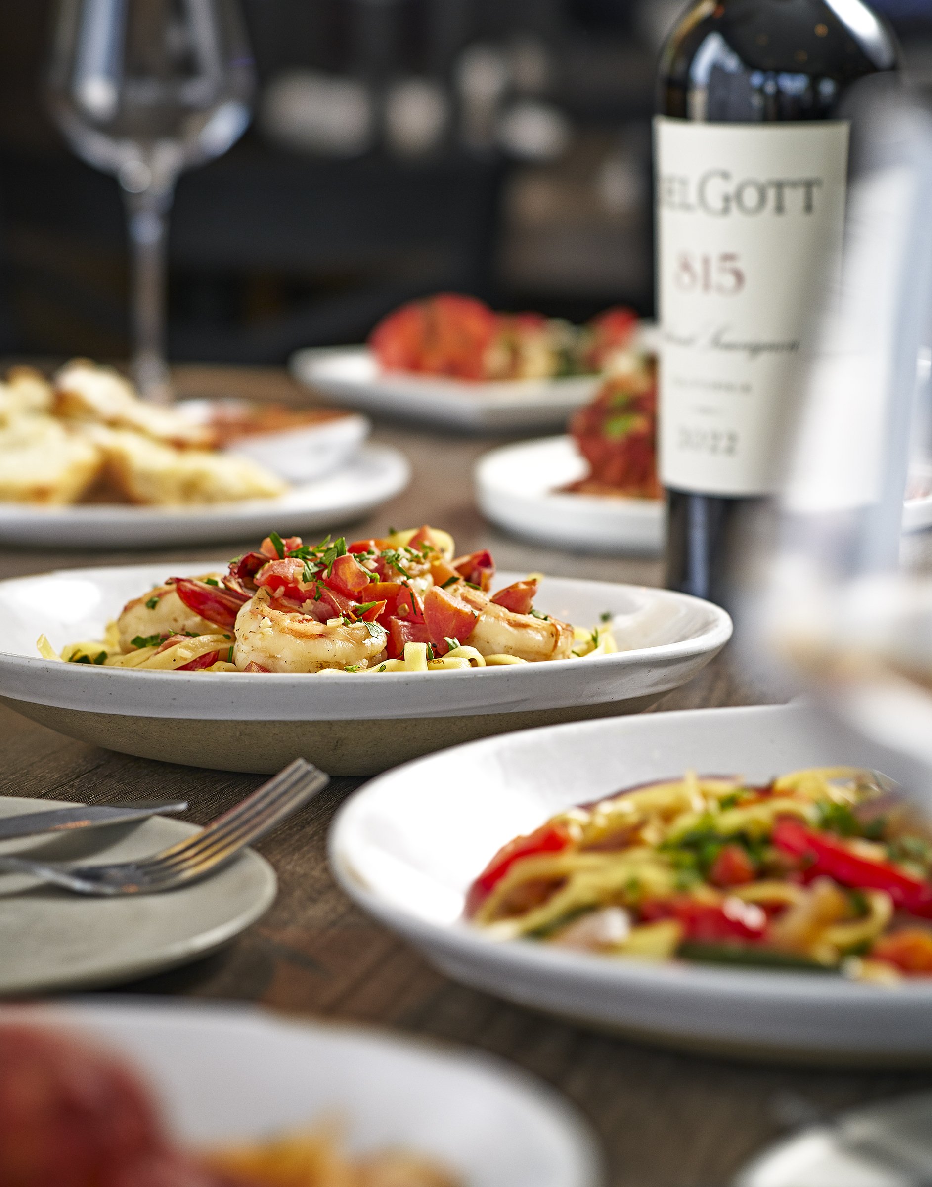Close-up of a plate of shrimp and pasta garnished with chopped green herbs, with other dishes and a table setting in the background.