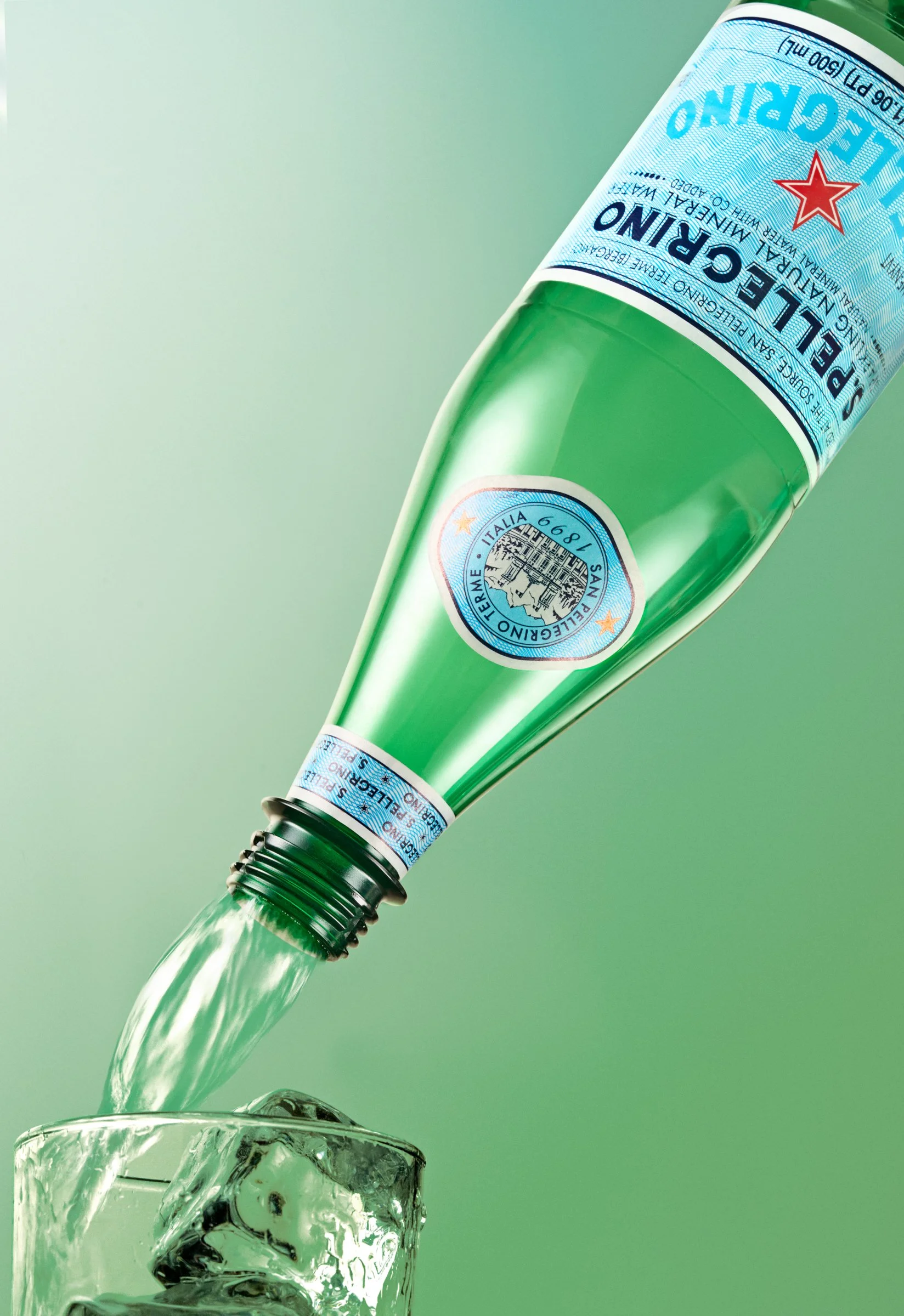 A green glass bottle of San Pellegrino mineral water pouring water into a glass with ice cubes on a light green surface.
