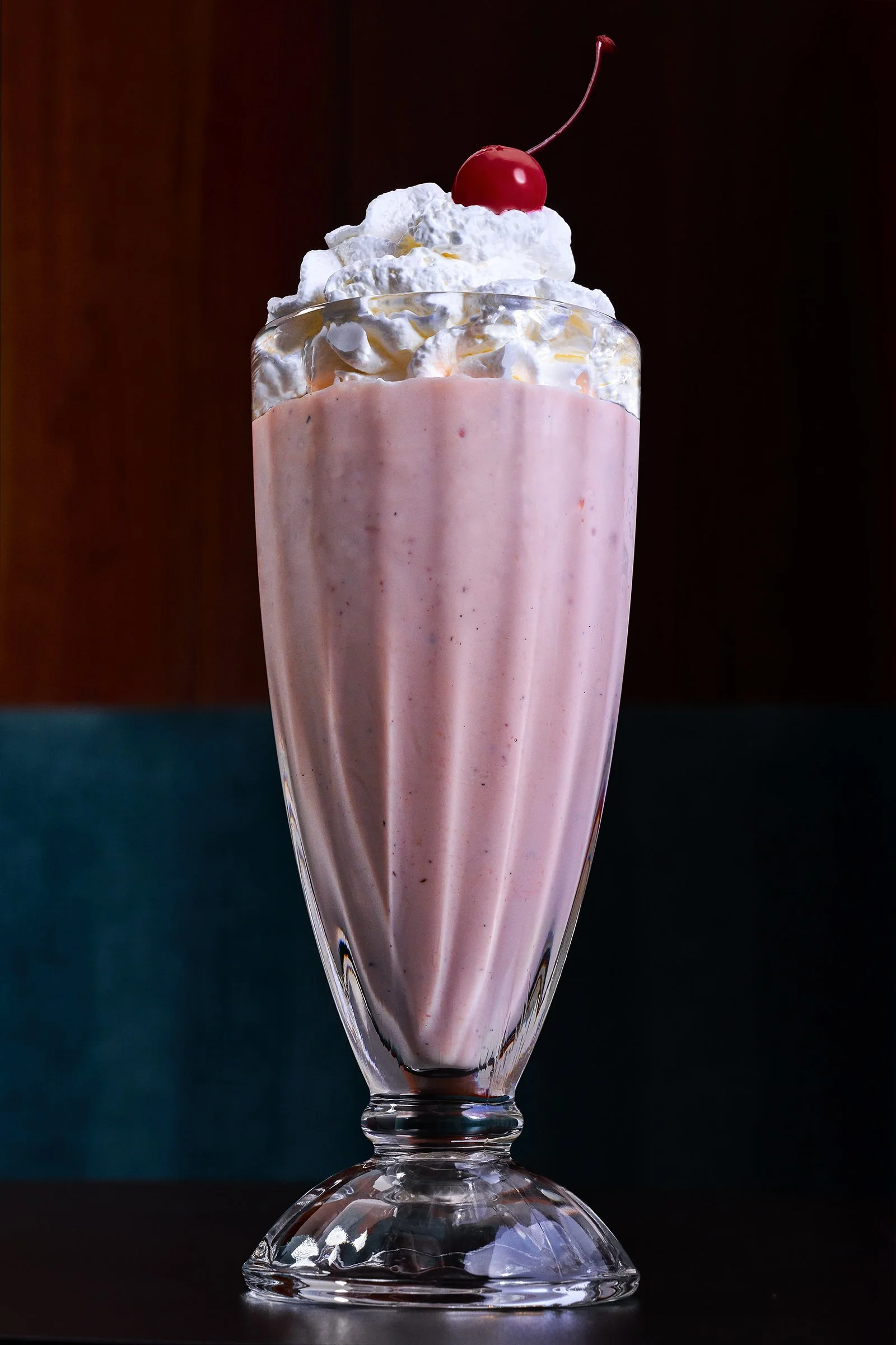 A tall glass with a pink milkshake topped with whipped cream and a cherry inside a tall glass with a wide base.