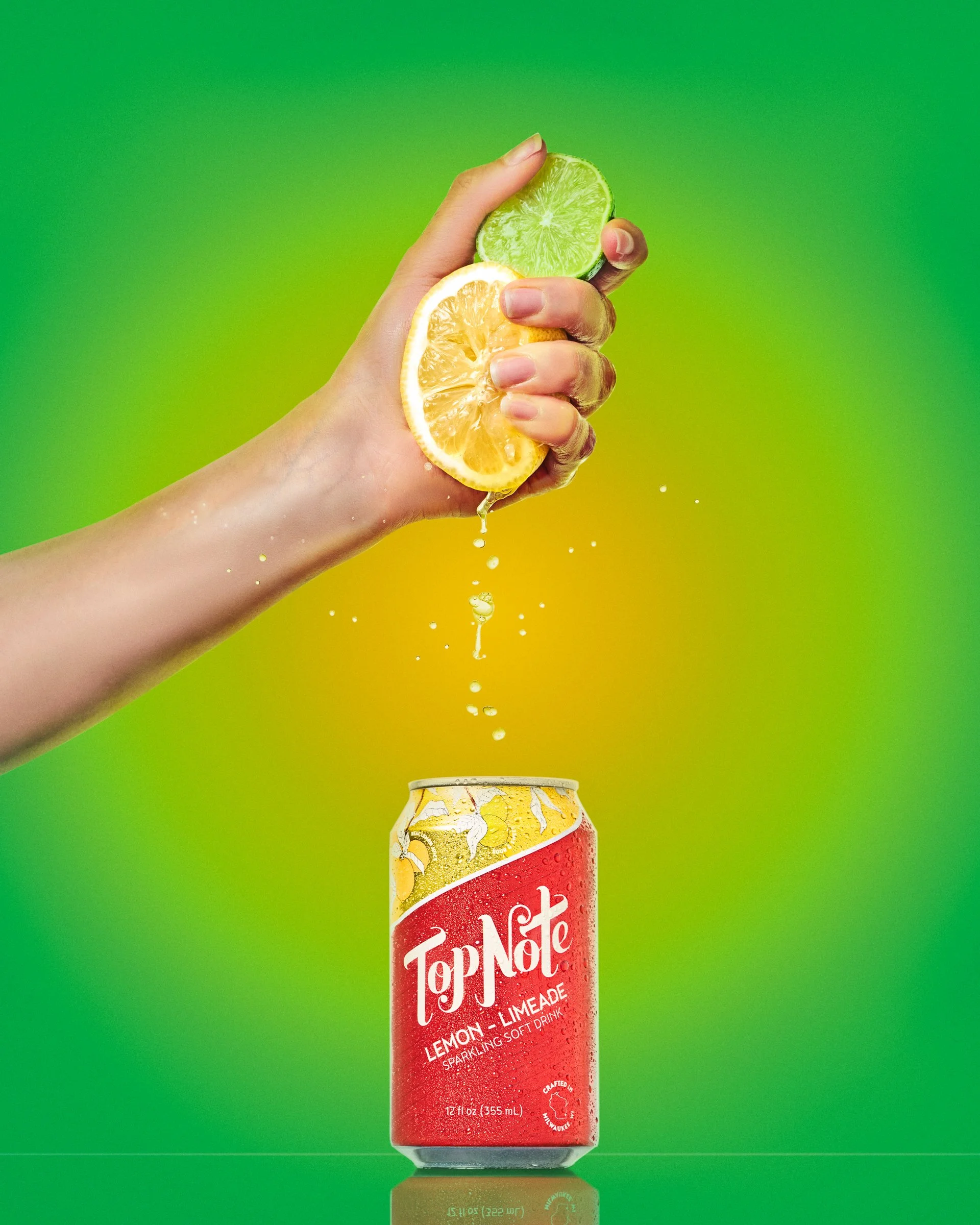 A hand squeezing a halved lemon and lime over a can of Top Note Lemon Limeade with drops of juice falling, set against a green background.