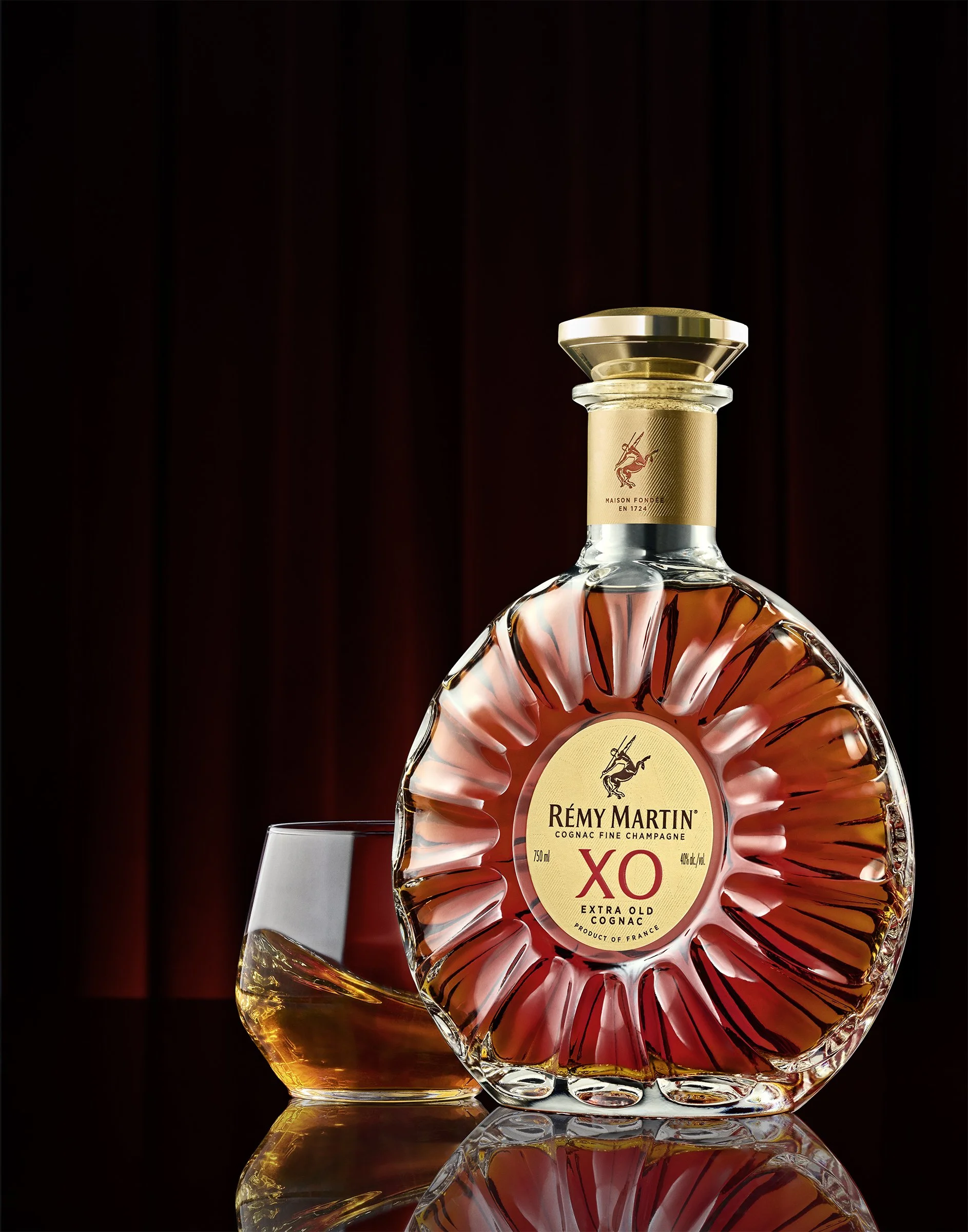 A bottle of Rémy Martin XO Extra Old Cognac with a glass of cognac beside it, set against a dark background.