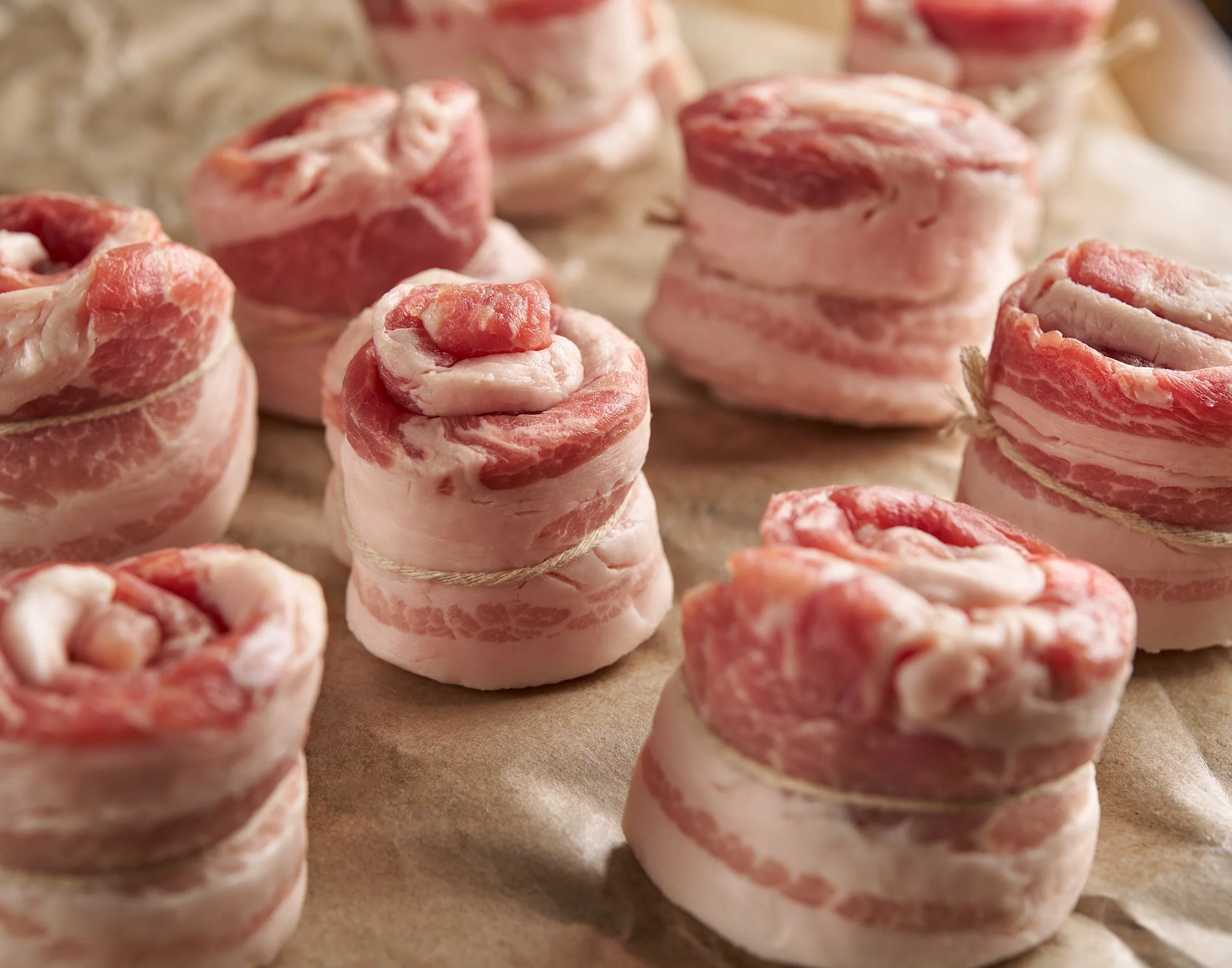 Close-up of small, round pieces of uncooked bacon-wrapped pinwheels on a brown surface.