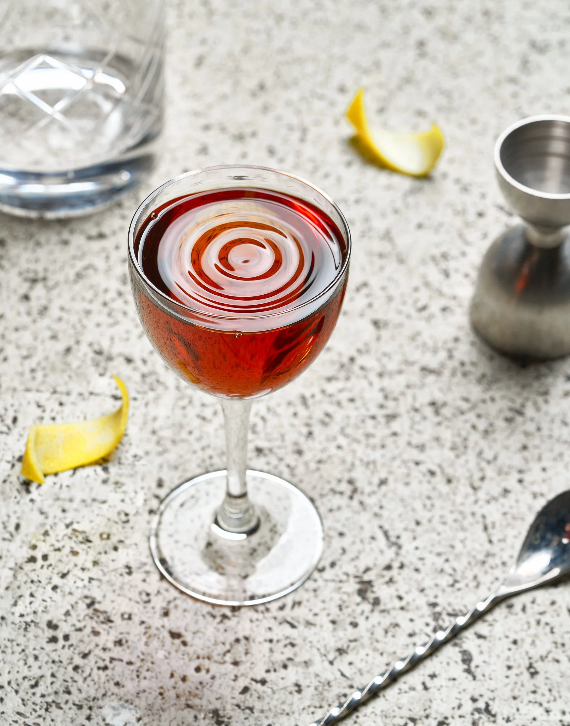 A glass of red wine on a speckled countertop with a lemon wedge, a cocktail shaker, a citrus squeezer, and a jigger in the background.