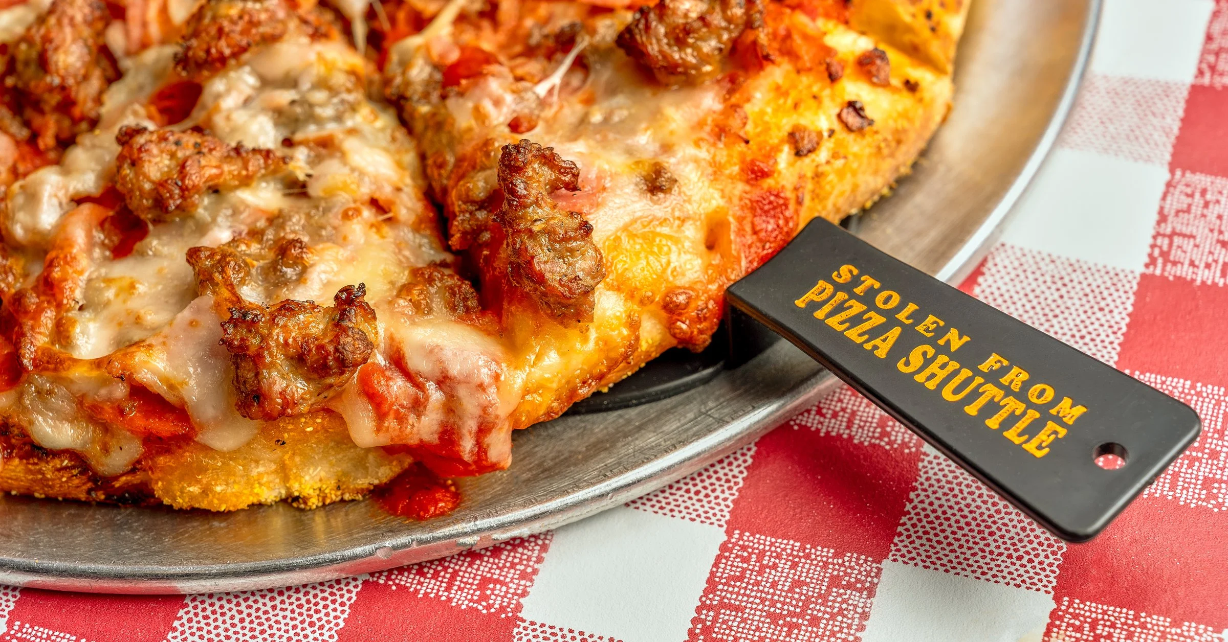 Close-up of a slice of meat lover's pizza with cheese, sausage, and ground beef on a metal tray with a black pizza cutter that has yellow text reading "Stolen from Pizza Shuttle"