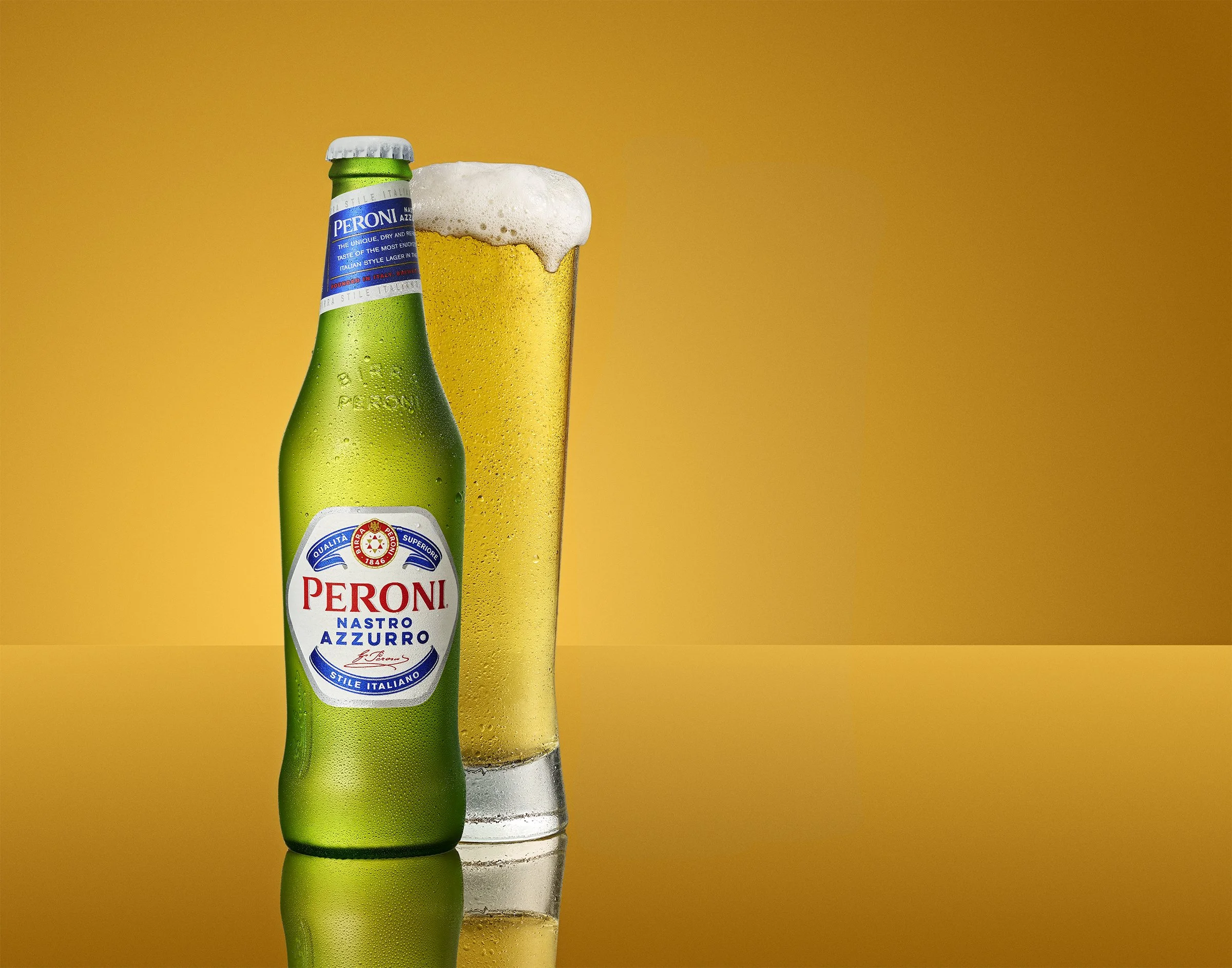 A green Peroni beer bottle with condensation next to a tall glass of beer with foam, both placed on a reflective surface against a yellow background.