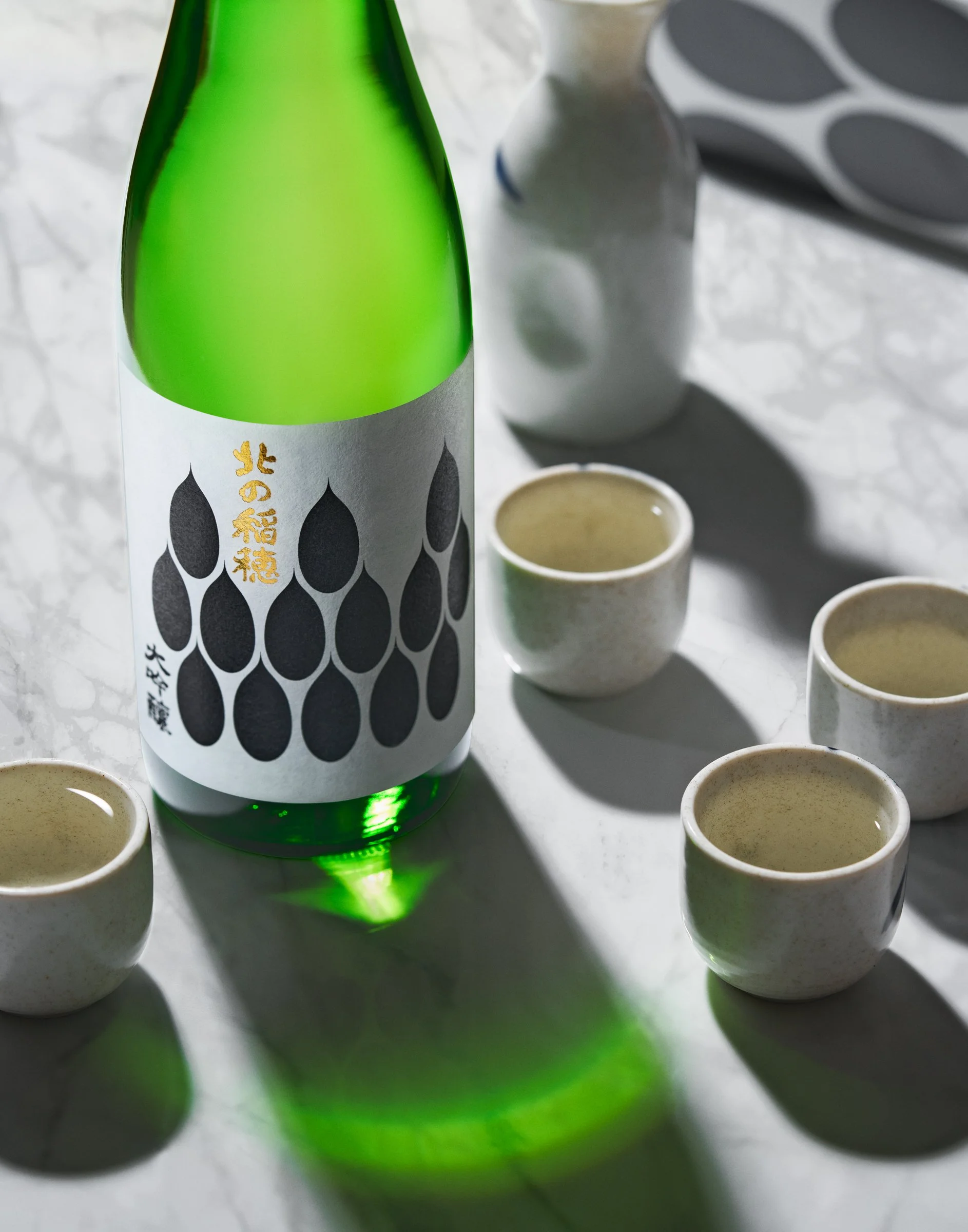 A green bottle of sake with Japanese characters on the label, surrounded by small sake cups on a white marble surface.