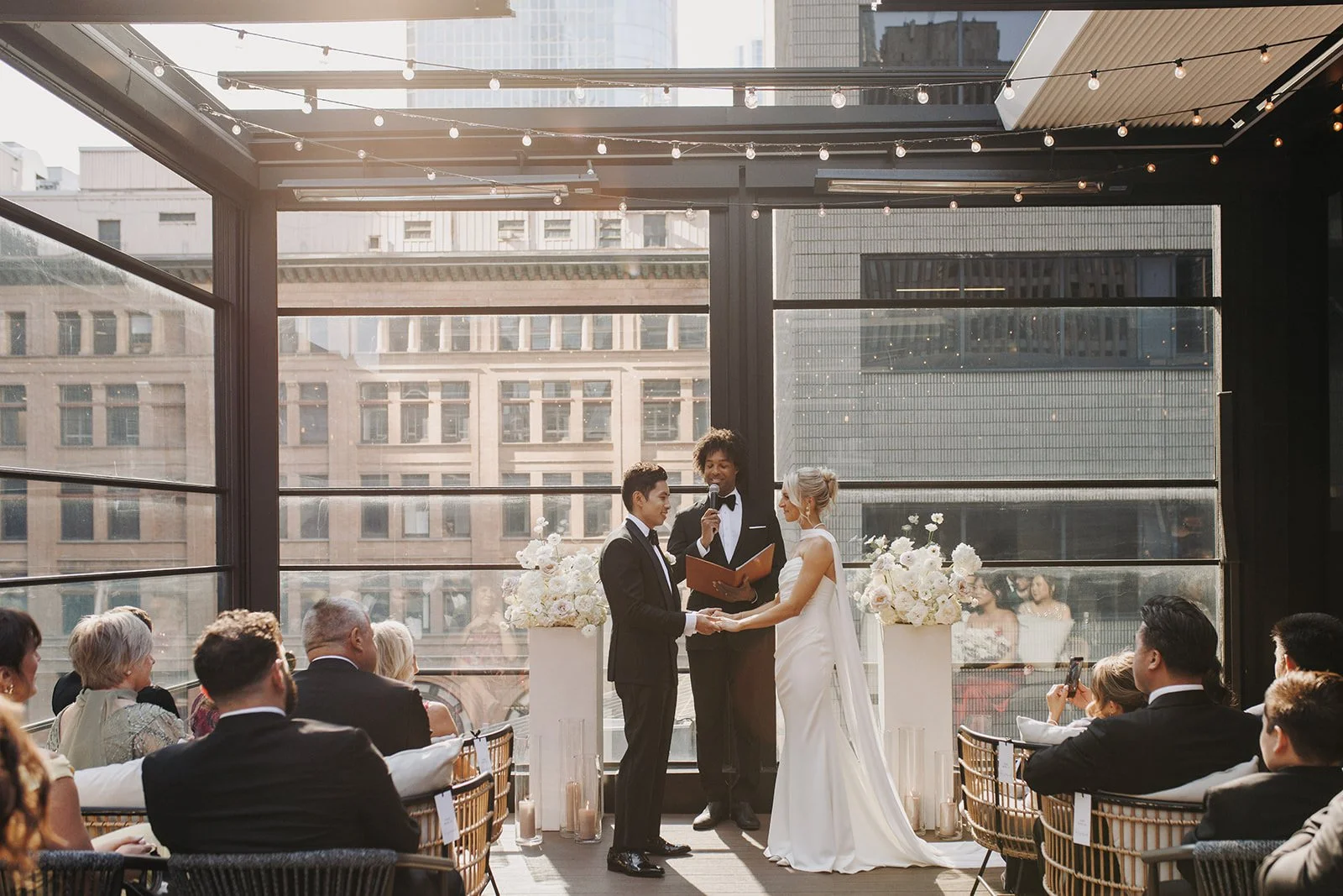A wedding ceremony with a couple holding hands in front of an officiant in a bright indoor venue with large windows and city buildings outside. Guests are seated on either side, dressed formally, observing the ceremony with Officiant Oren Williamson