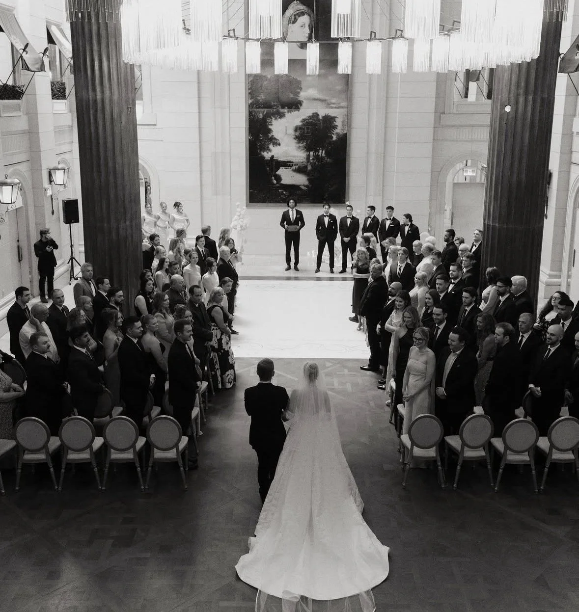 Black and white photo of a wedding ceremony in a grand hall with many guests seated on either side. The bride in a long dress with a veil and the groom in a tuxedo walk down the aisle toward a group of groomsmen at the front.