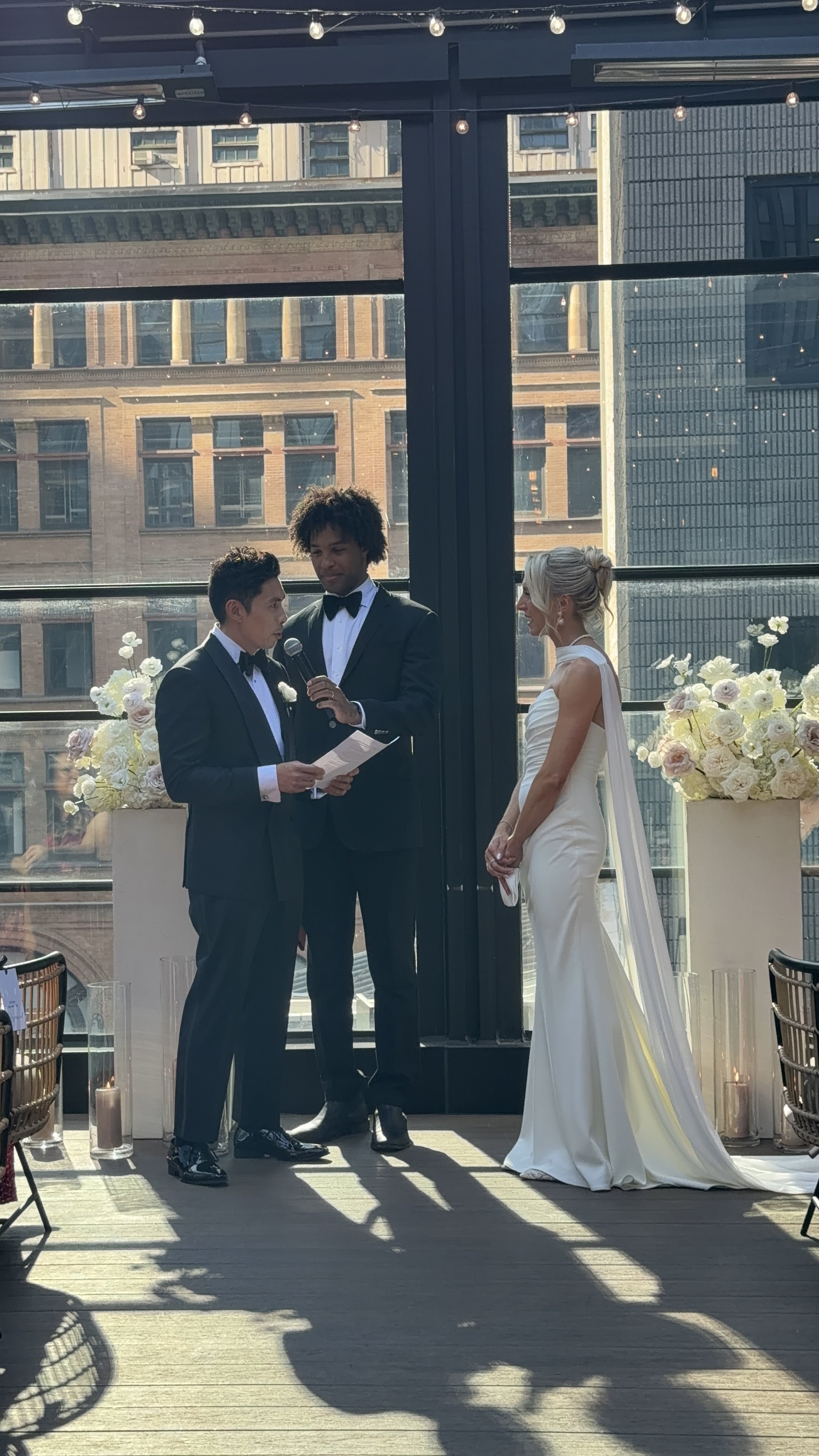 A wedding ceremony taking place indoors with city buildings visible through large windows. A man in a tuxedo is reading vows from a paper while another man in a tuxedo holds a microphone. A woman in a white wedding gown stands facing them, smiling. Flowers and candles are arranged on either side of the ceremony area.