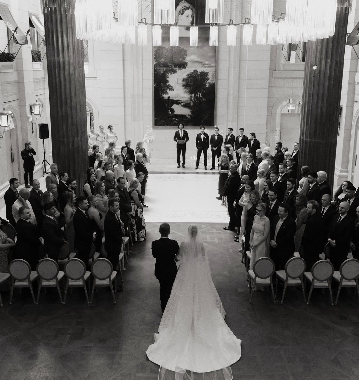 A bride and groom facing each other at the altar in a wedding ceremony, with guests seated on either side of the aisle in a grand hall decorated for a wedding officiant Oren Williamson