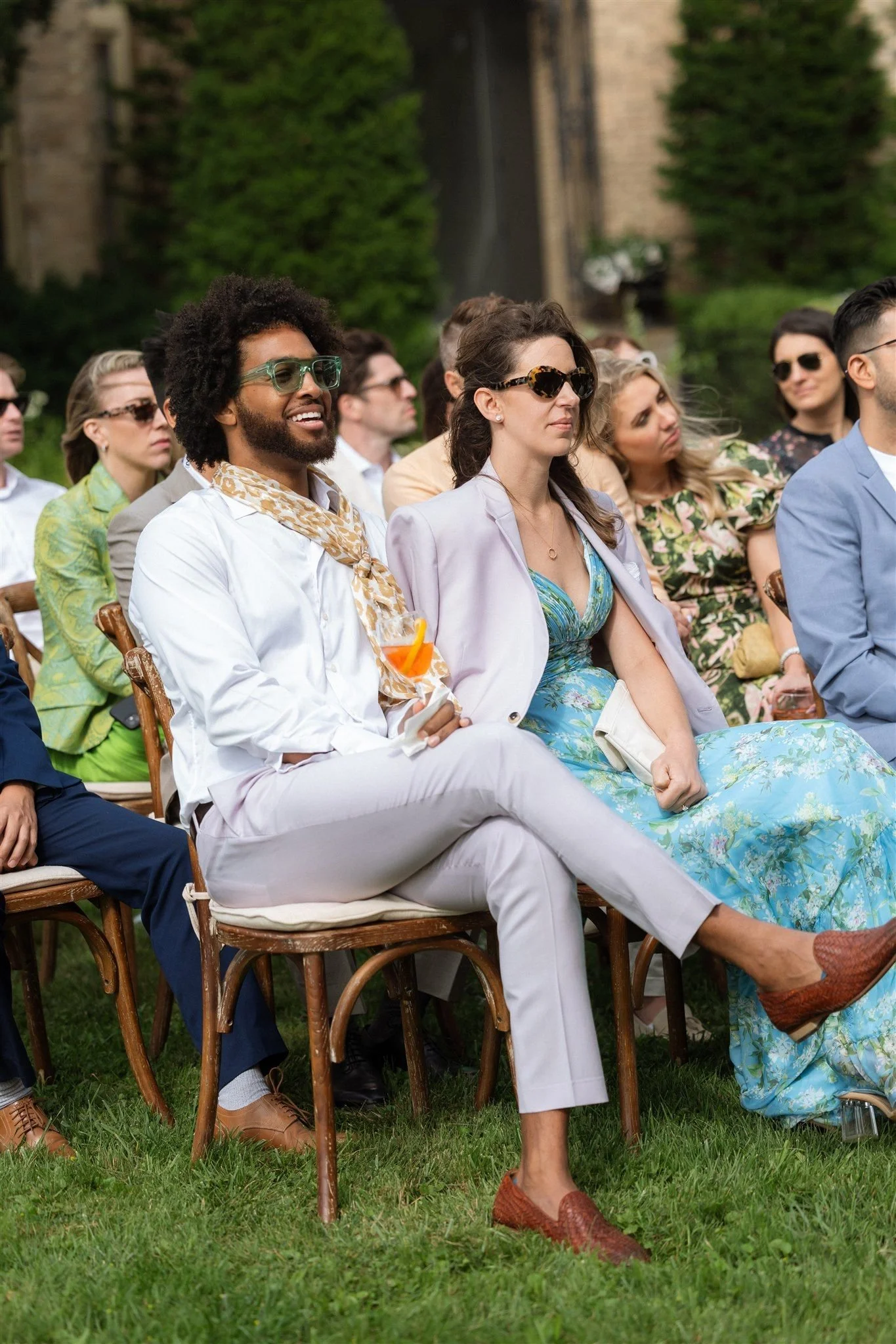 A group of people attending an outdoor event, seated on wooden chairs on grass, dressed in colorful summer clothing, many wearing sunglasses, with trees and a stone structure in the background.