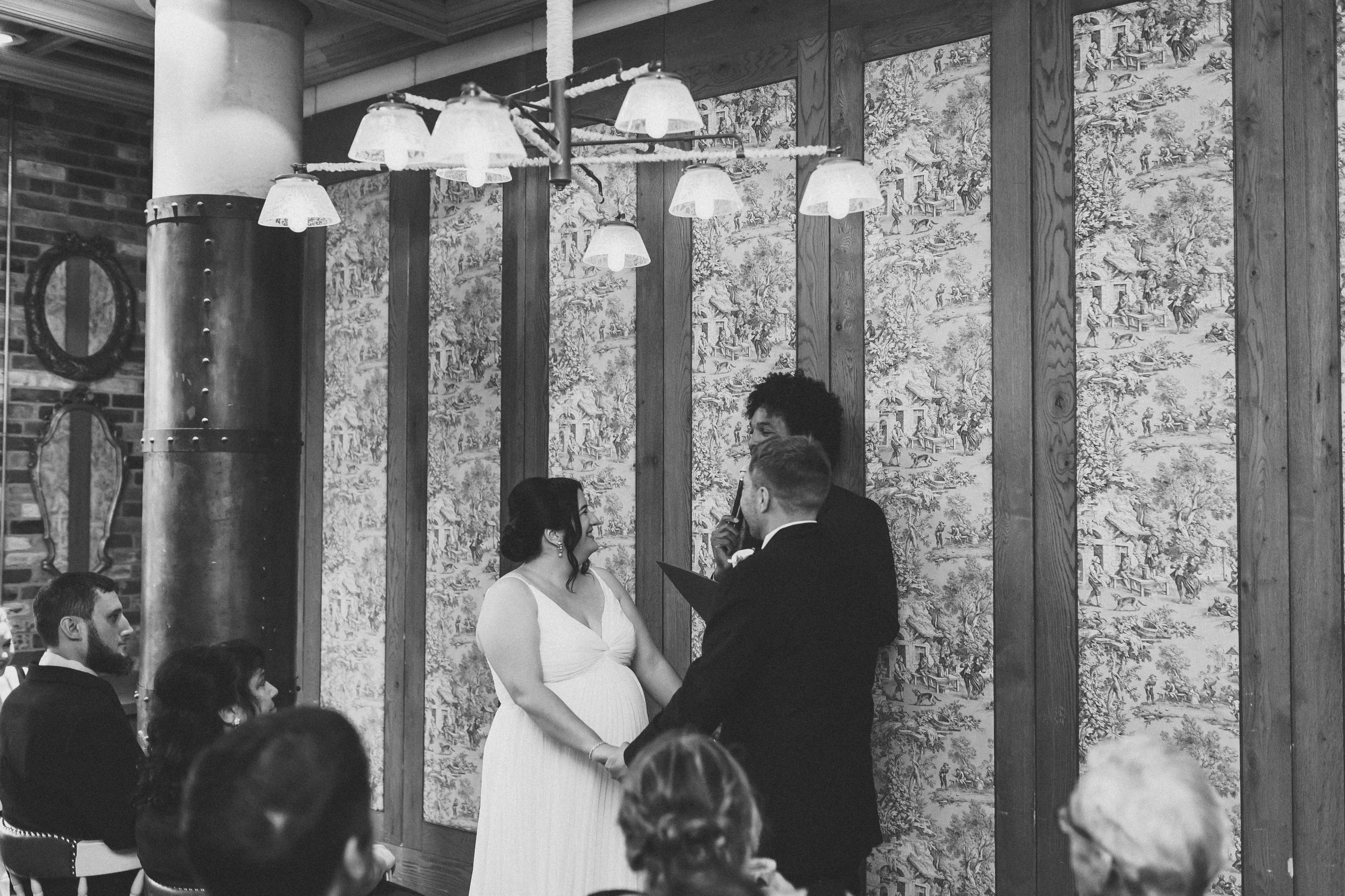 A black and white photo of a wedding ceremony where a couple is exchanging vows, with people seated around observing. The room has vintage wallpaper and hanging lights.