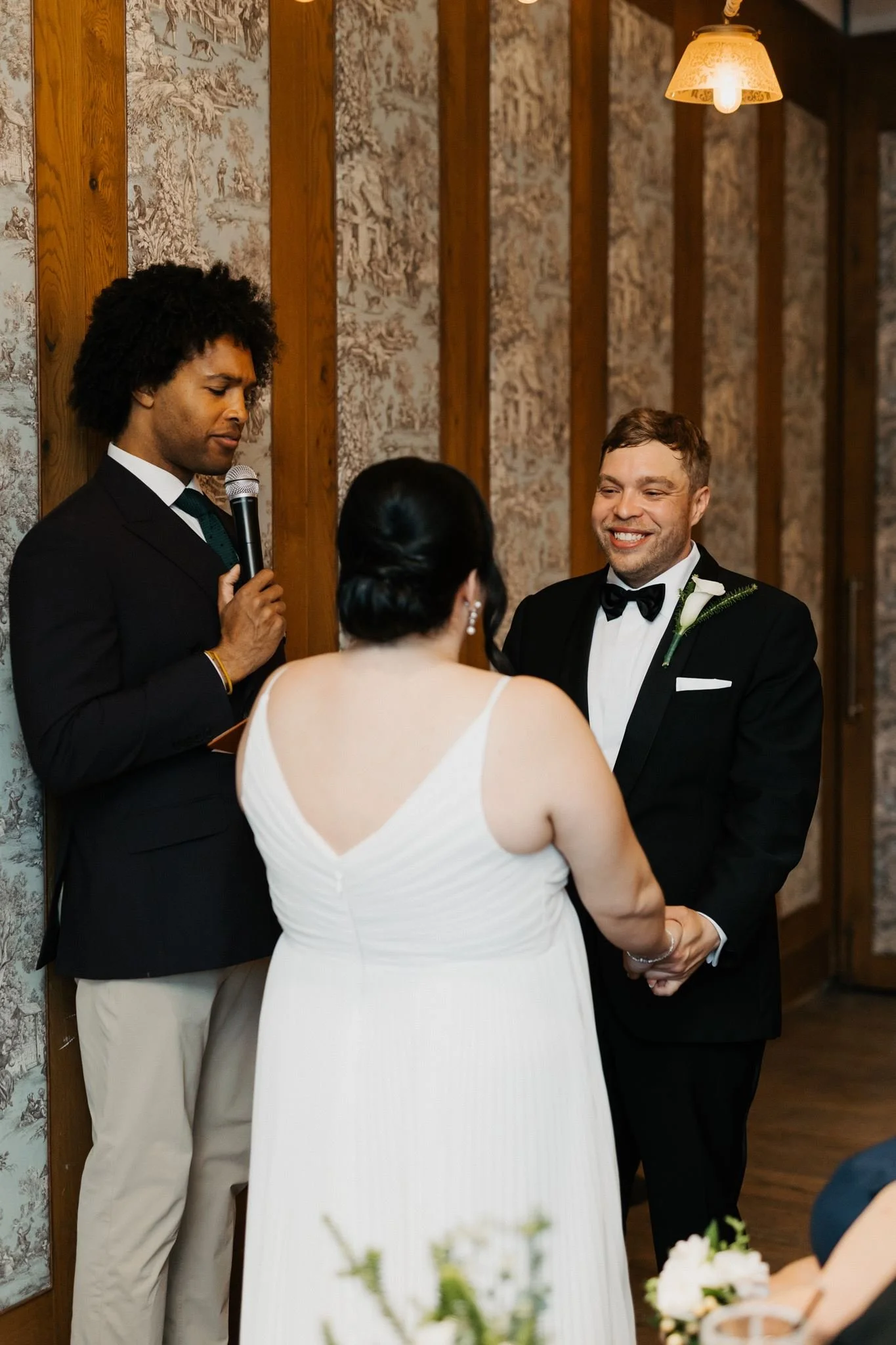 A wedding ceremony with a bride and groom holding hands, facing an officiant with a microphone, in a warmly lit room with wooden accents and patterned wallpaper.
