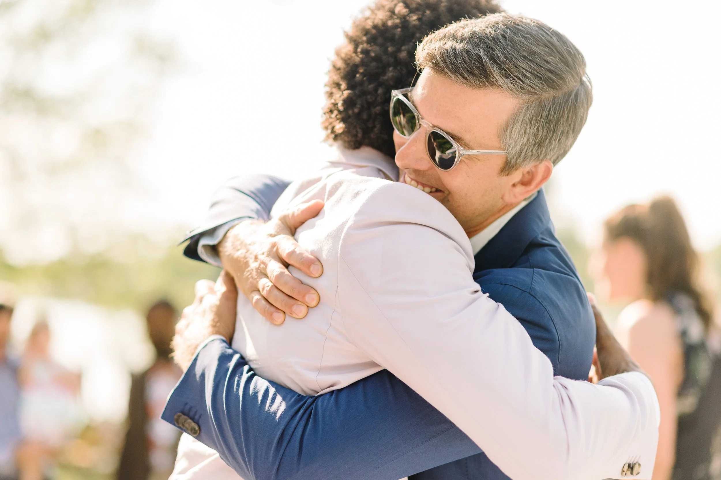 Two people hugging outdoors during daytime, smiling, with other blurred people in the background.
