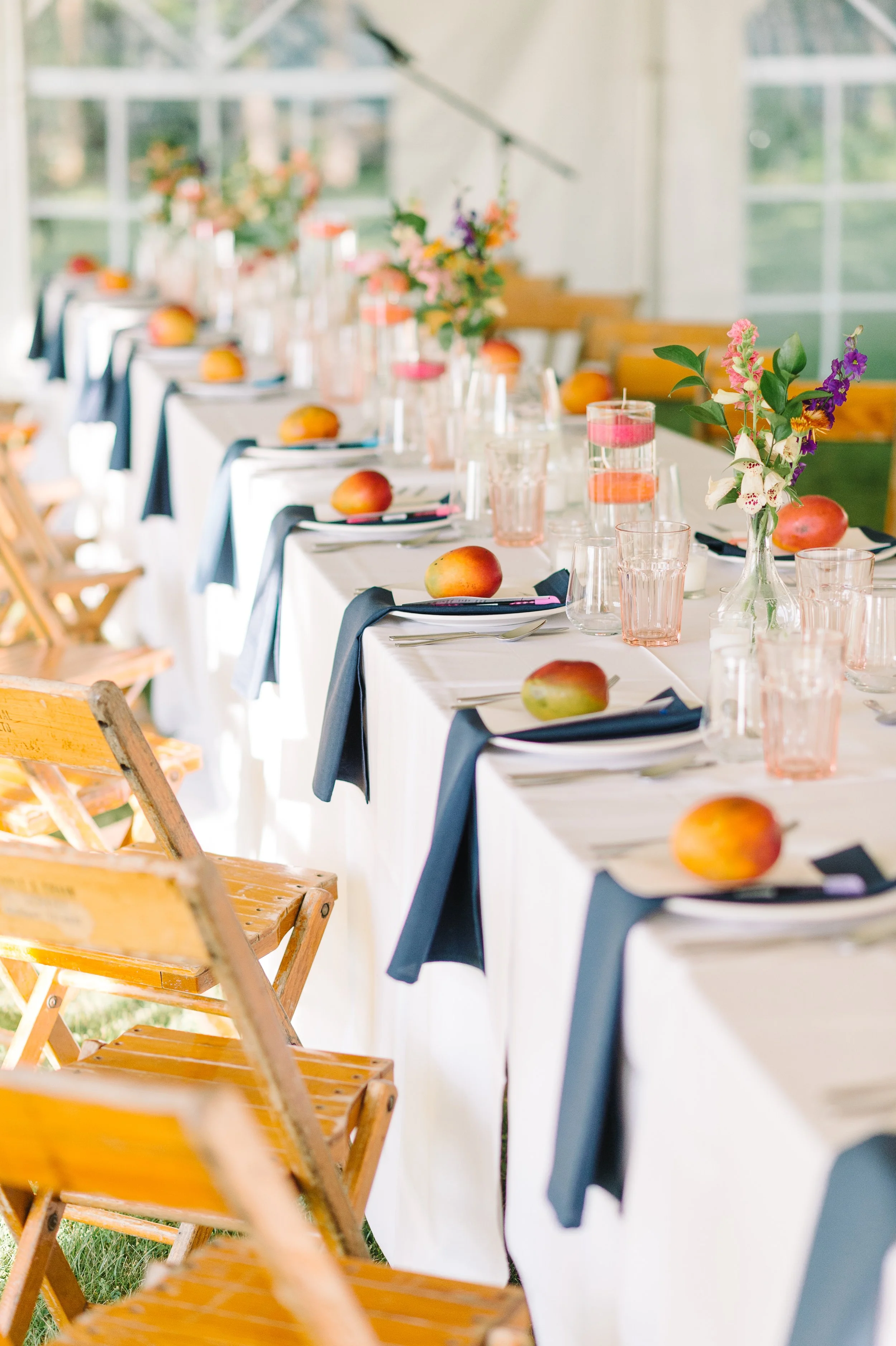 A long banquet table decorated with flowers and mangoes, with wooden chairs on both sides, set in a bright room with large windows.