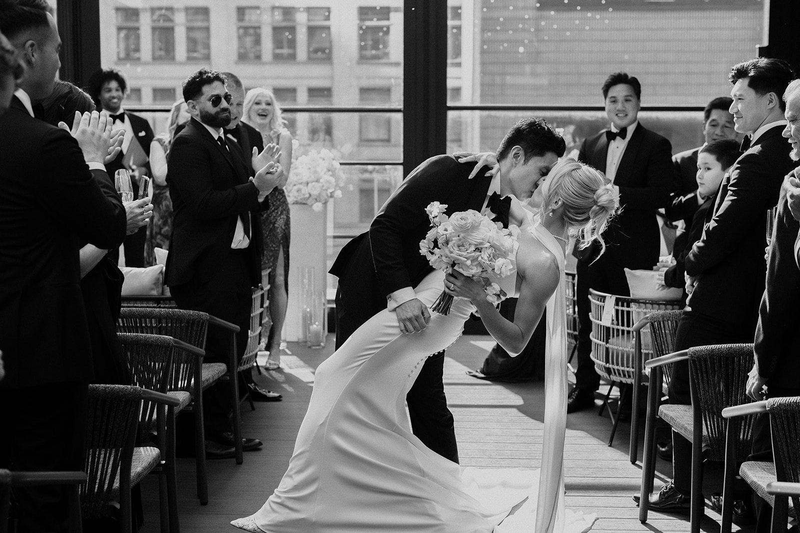 A black-and-white photograph of a wedding ceremony. The groom is leaning over and kissing the bride, who is holding a bouquet of flowers. Guests are standing and clapping on either side of the aisle, with large windows and a cityscape visible in the background.