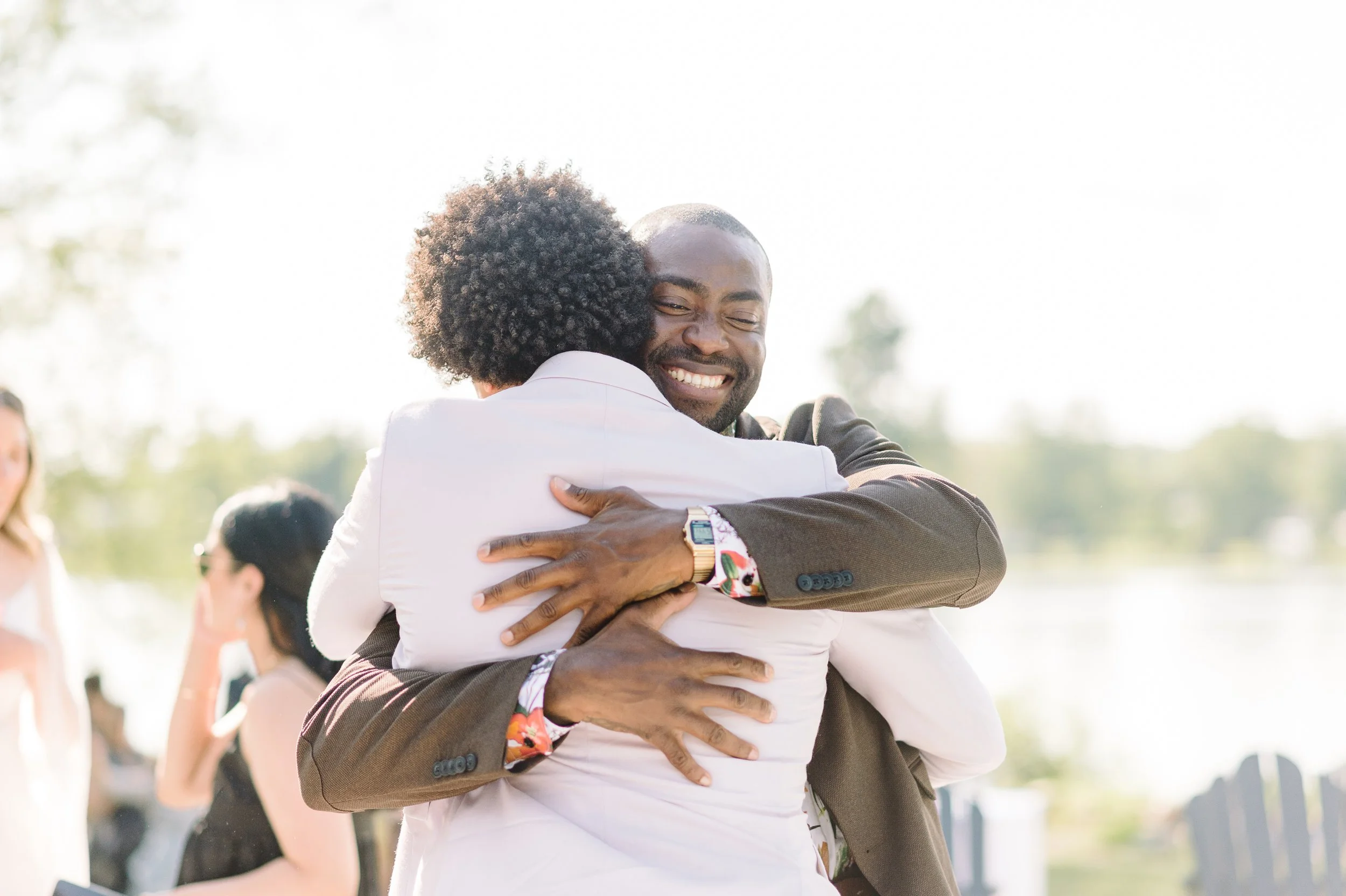 Two people hugging outdoors on a sunny day, with other people in the background.