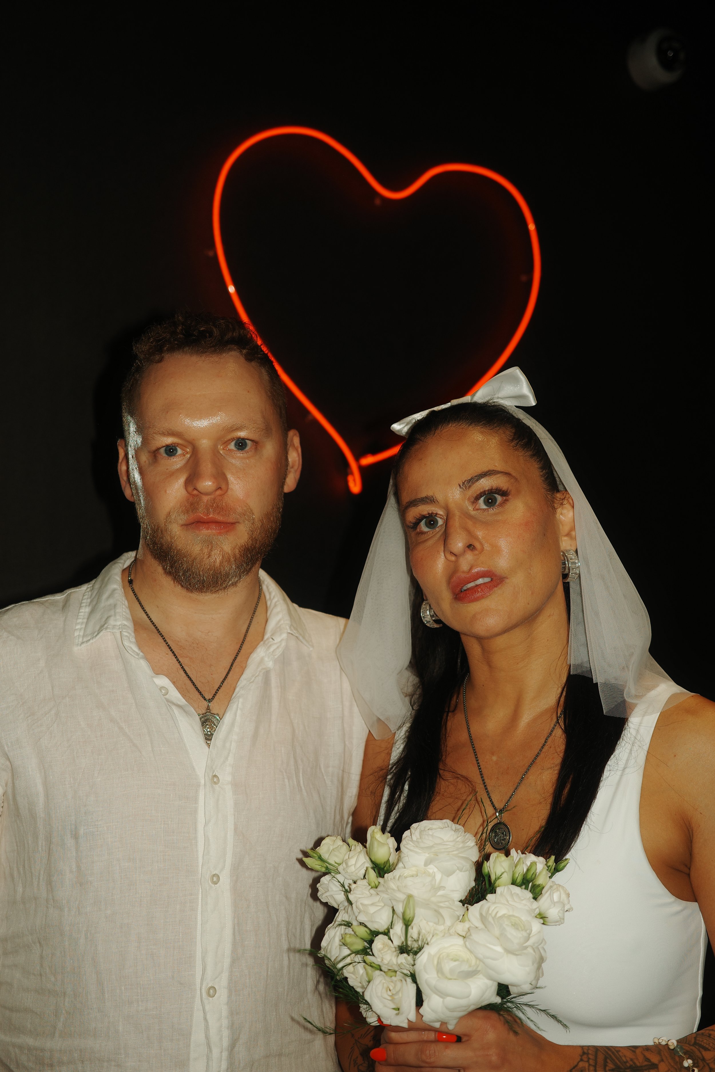 A man and woman dressed in wedding attire stand close together in front of a black background with a red neon heart above their heads. The woman is holding a bouquet of white flowers, wearing a white dress and a veil, and the man is wearing a white shirt with a necklace. The woman has dark hair and makeup, and the man has short hair and a beard.
