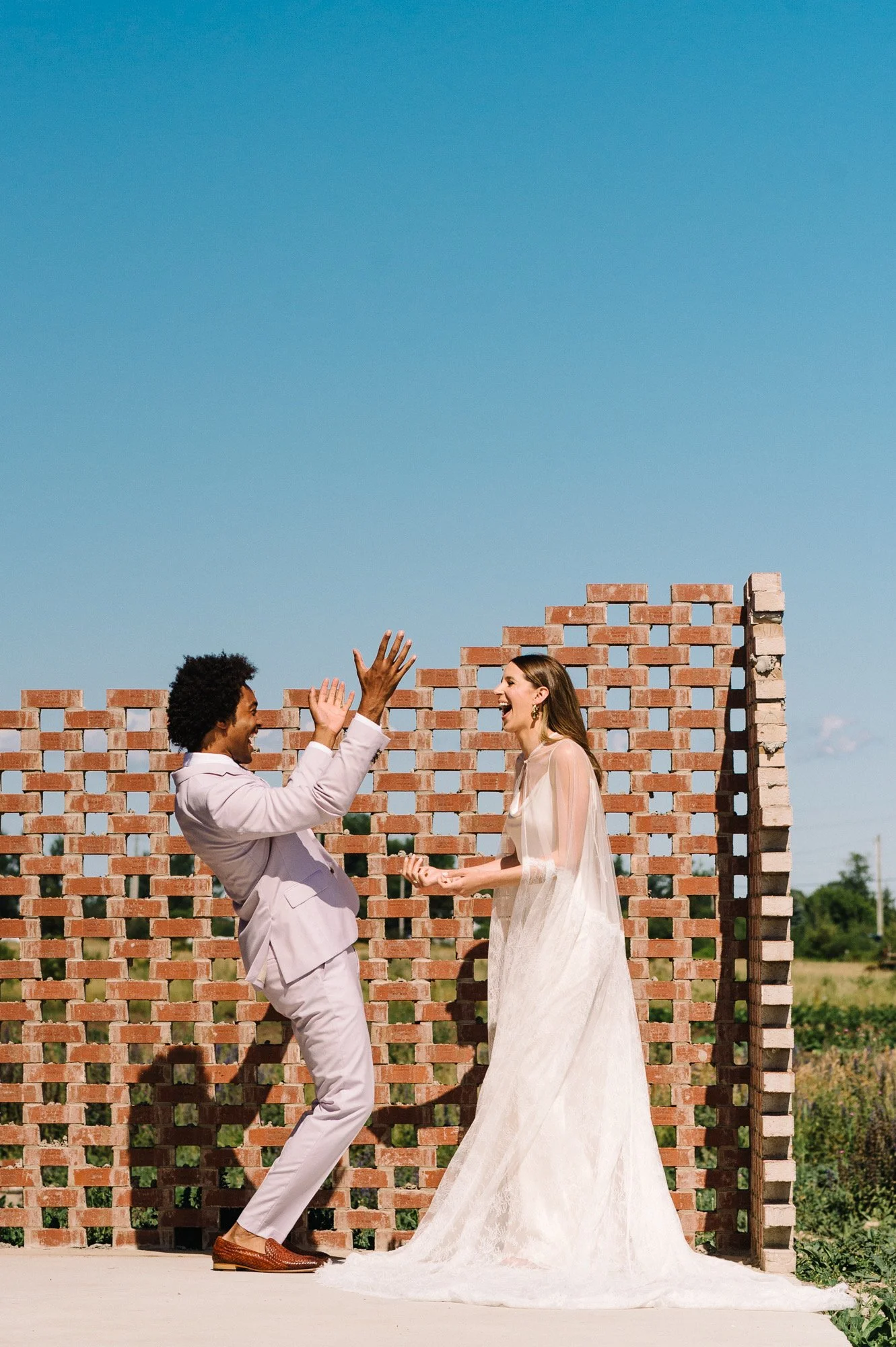 A man and woman dressed in formal attire, high-fiving each other, standing outdoors in front of a partially built brick wall with a clear blue sky.