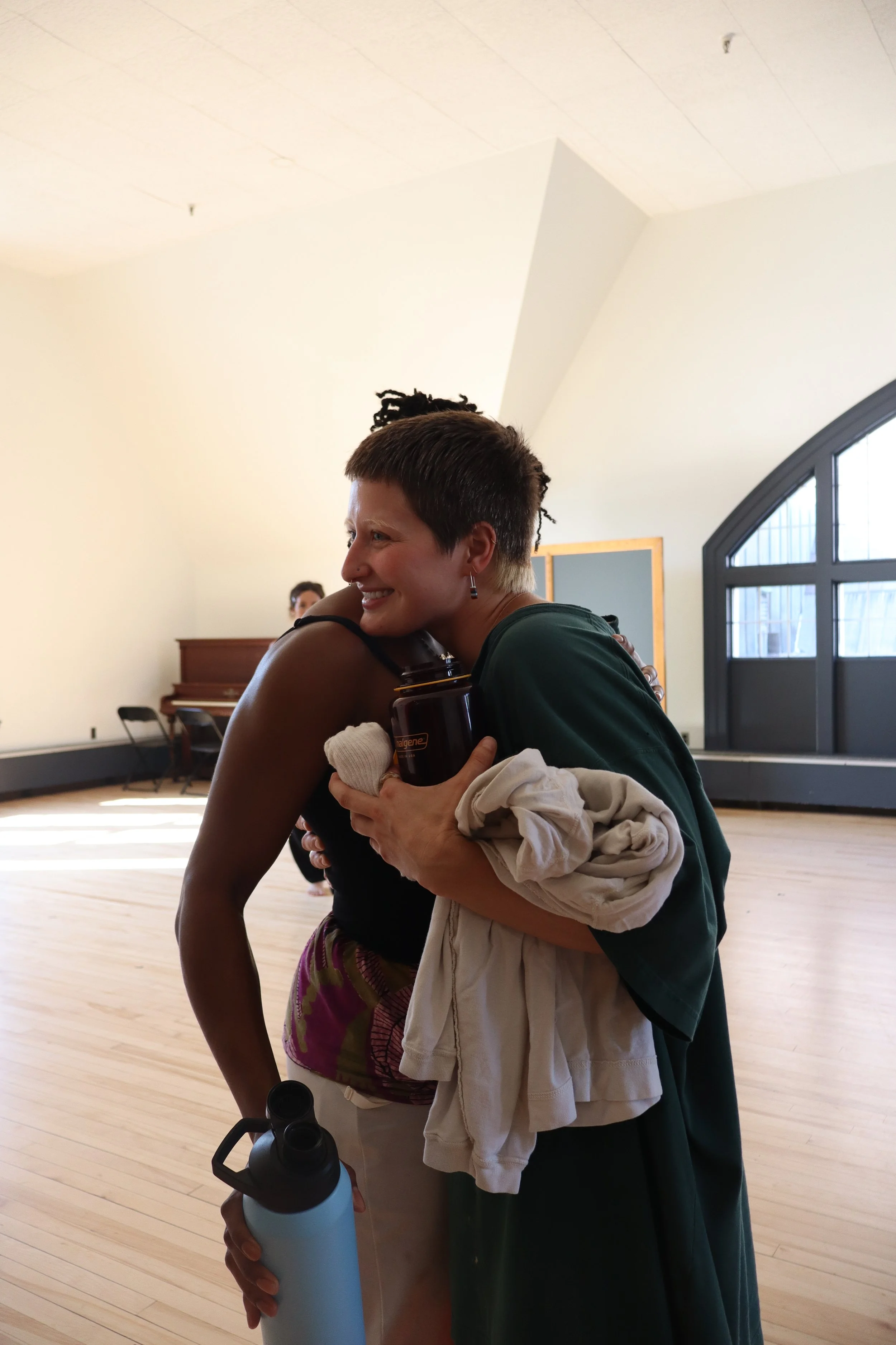 Two women hugging inside a bright room with wooden floors and high large windows. One woman has short hair and is holding a water bottle, the other is wearing a tank top and purple shorts. There is a piano against the wall and a black folding chair i