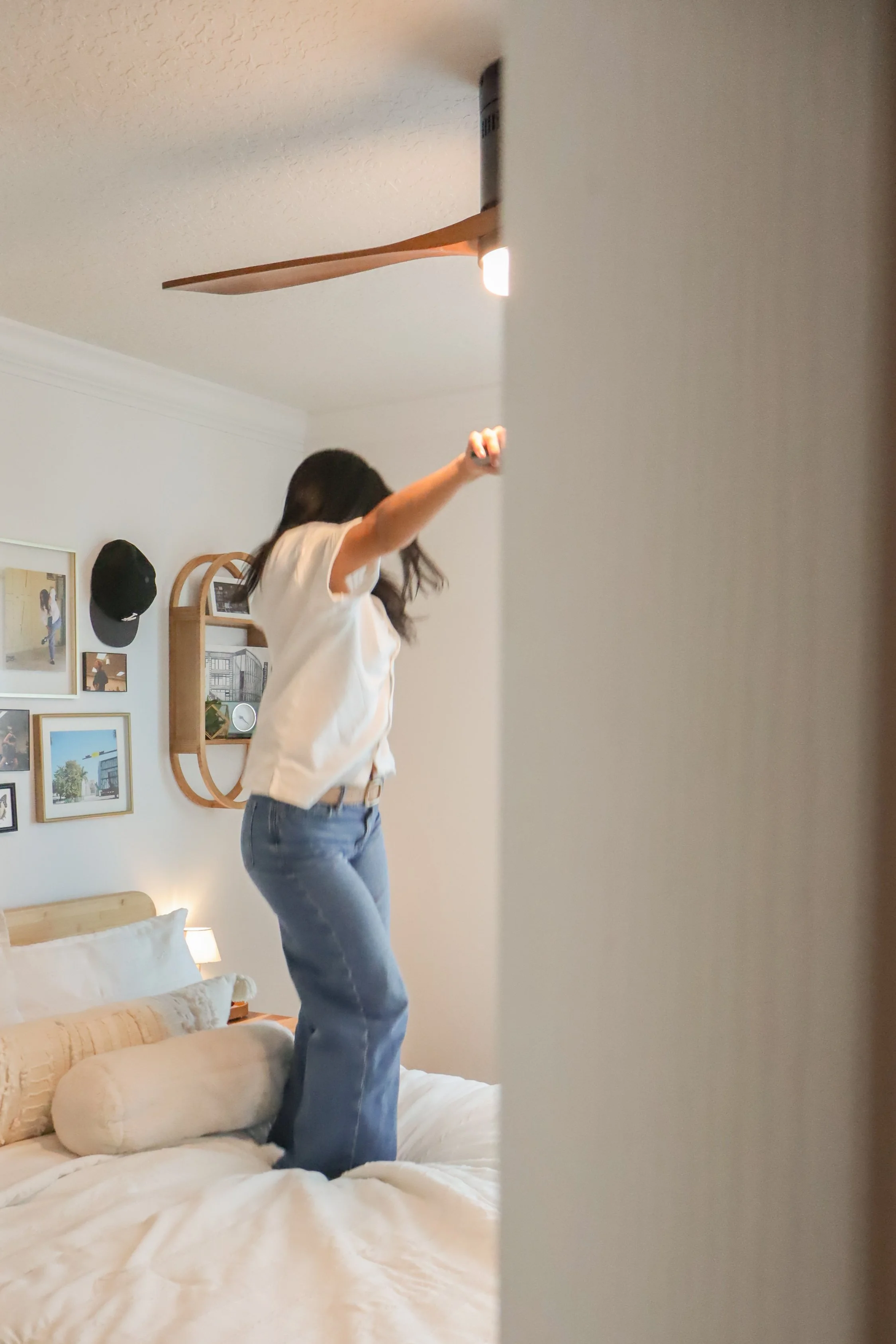 A woman with long dark hair wearing a white t-shirt and blue jeans standing on a bed, reaching up towards a ceiling fan in a bedroom decorated with framed pictures on the wall and a black baseball cap hanging on the wall.