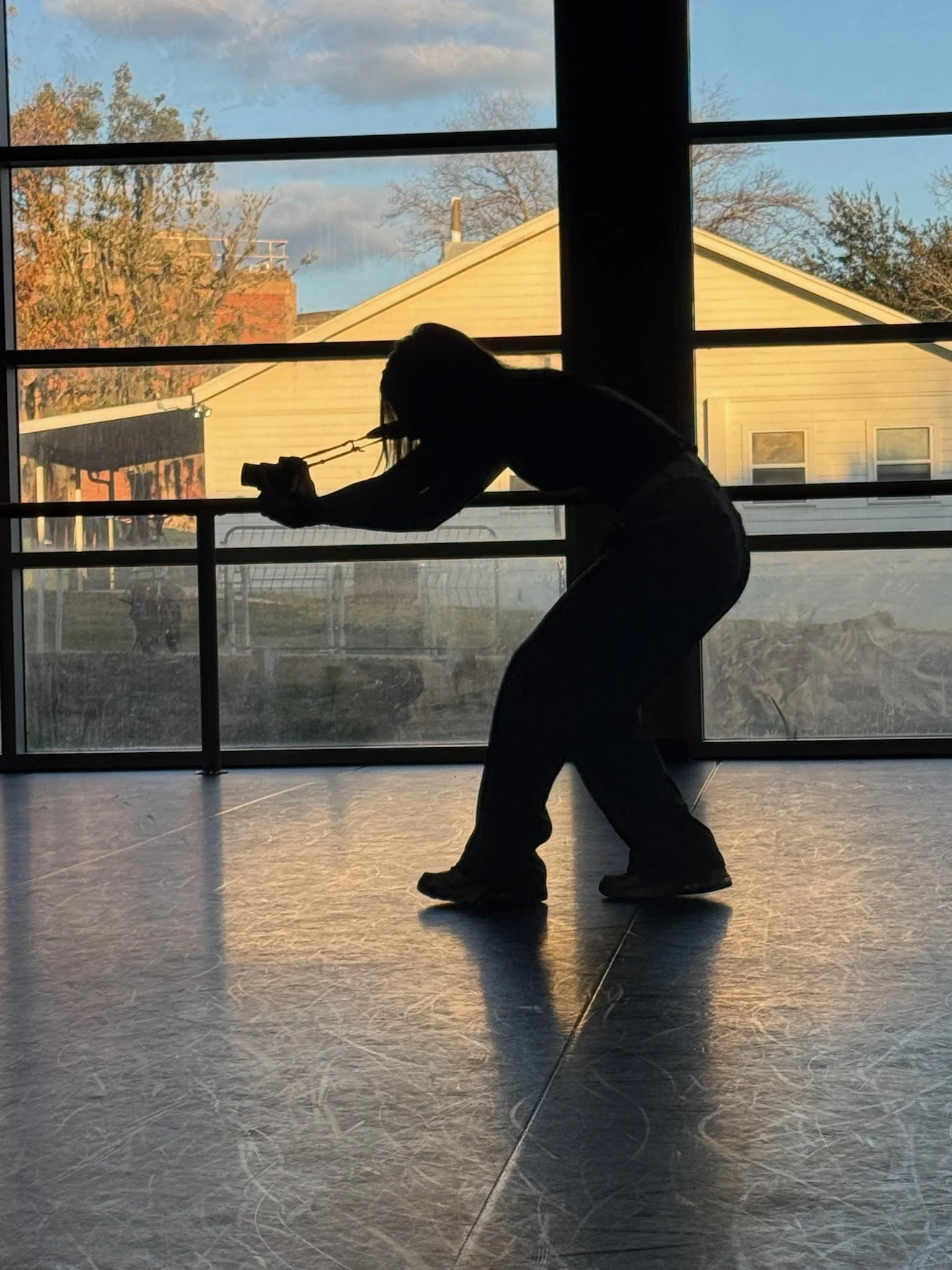 A person with long hair is taking a photo or video of themselves with a smartphone, in front of large windows showing a house and trees outside during daytime.