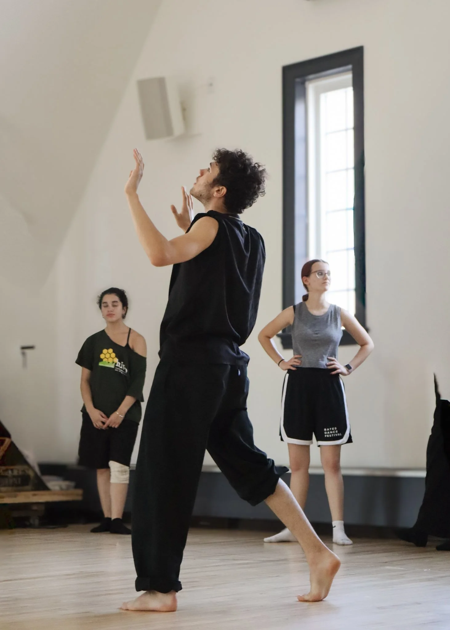 A group of dancers practicing in a dance studio, with a male dancer in the foreground demonstrating a dance move while others observe.