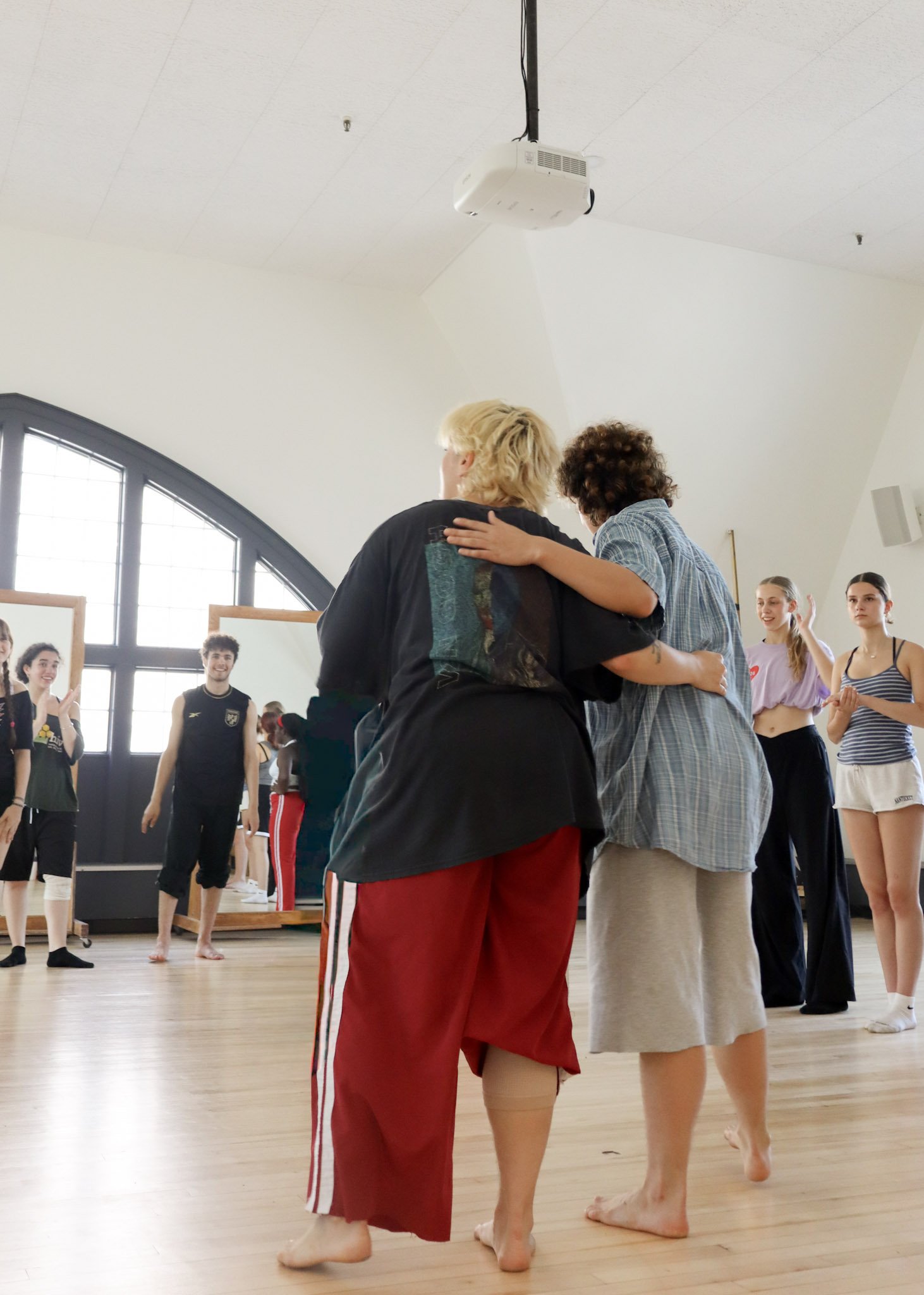 People standing in a circle barefoot in a spacious dance studio with large windows, some holding each other, others clapping or smiling.
