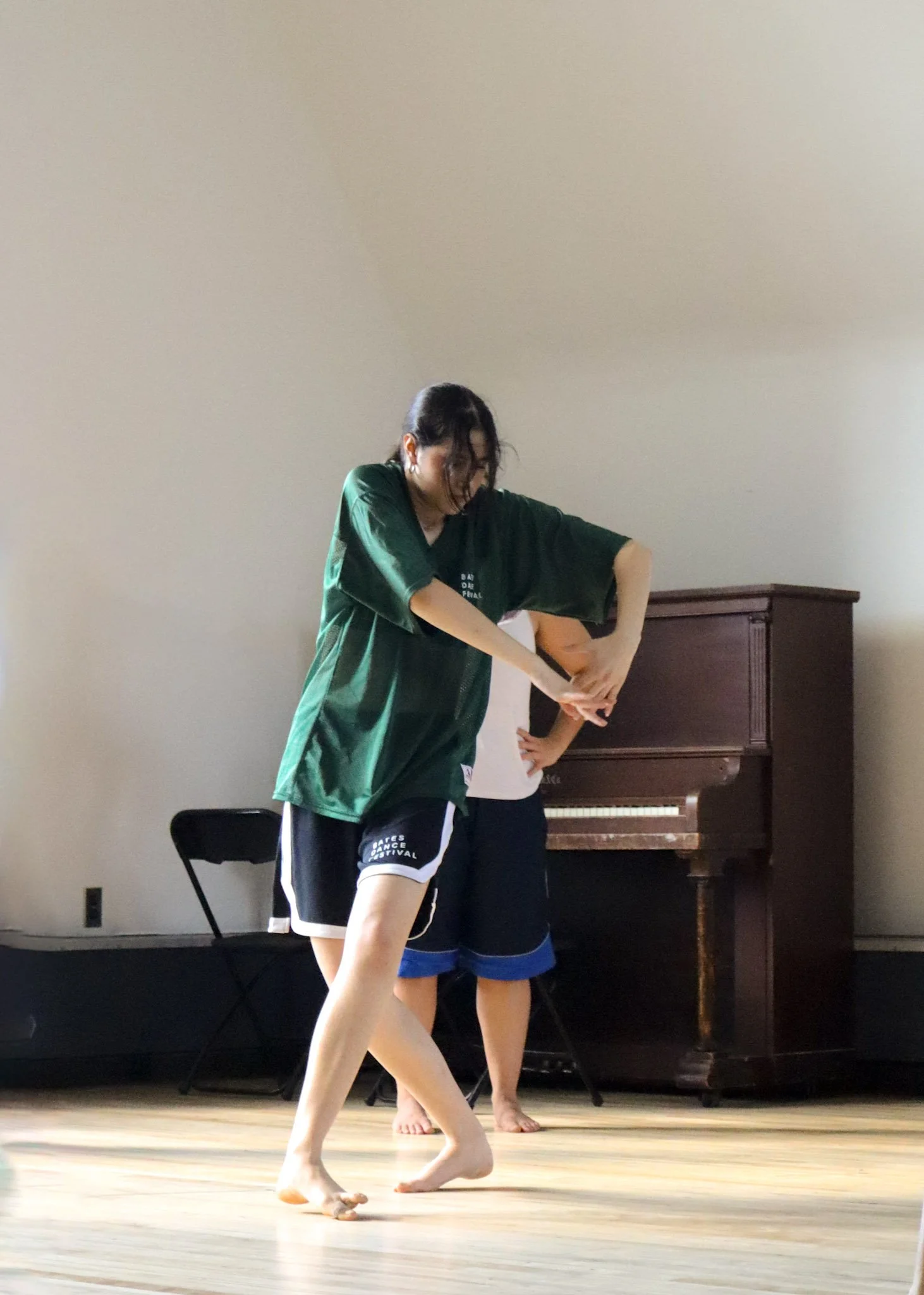 Two dancers practicing barefoot on a wooden floor, with a piano and a chair in the background.