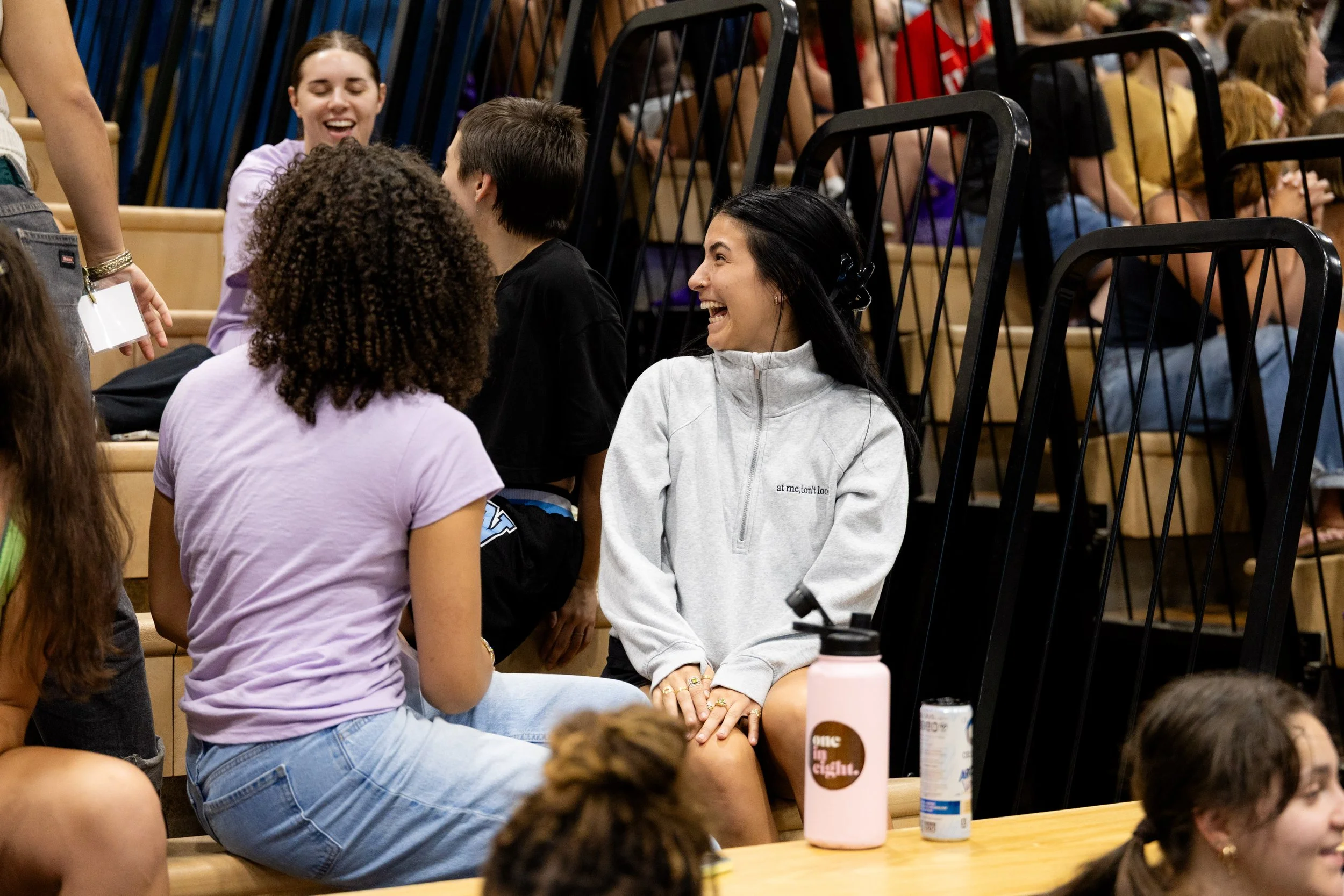 A group of people sitting on bleachers at an indoor event, engaging in conversation, smiling, and laughing. Some are standing, and others are seated, with beverages and personal items on the tables nearby.