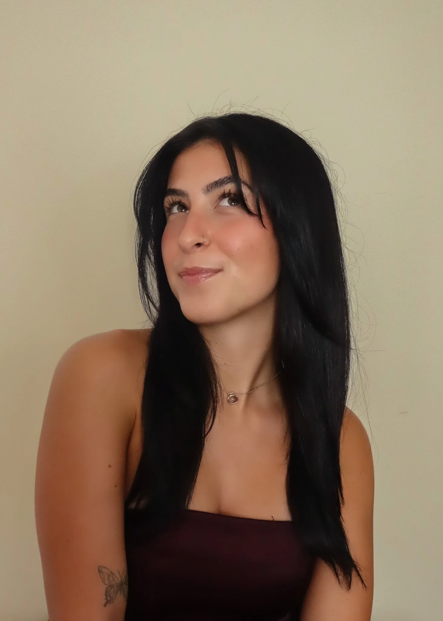 A young woman with black hair, a nose piercing, and a butterfly tattoo on her arm, wearing a strapless top and a necklace with a circular pendant, smiling and looking to the side against a plain wall.