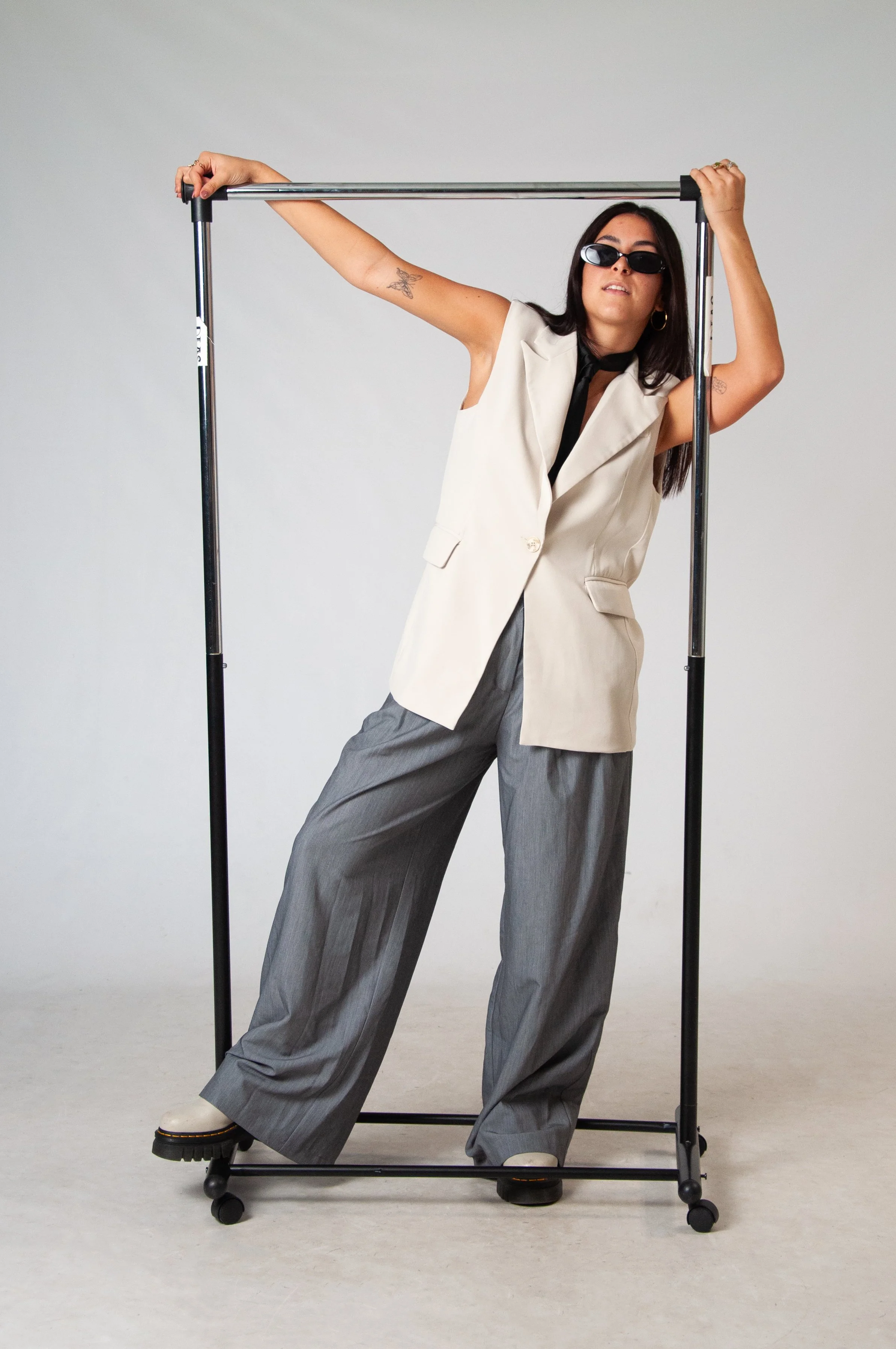 A woman wearing sunglasses, a sleeveless beige blazer, gray wide-leg trousers, and black shoes stands inside a white studio, leaning against a black metal clothing rack, with one arm resting on the top bar and one arm holding the rack for support.