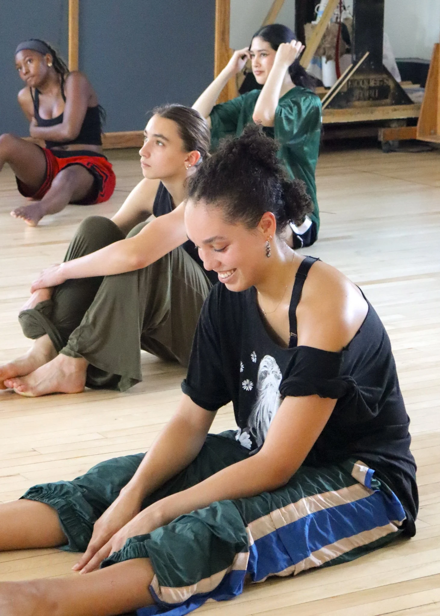 Four young women sitting on a wooden floor, stretching or relaxing in a dance or yoga class. They are seated with their legs extended and leaned forward, smiling and focusing on their stretches.