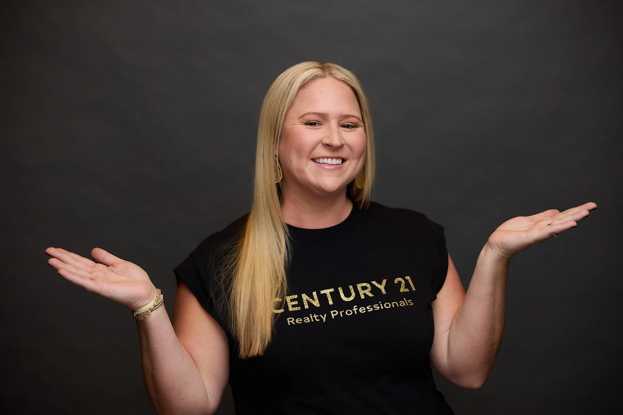 A young woman with long blonde hair smiling and shrugging her shoulders with her hands outward, wearing a black T-shirt with gold lettering and gold earrings, against a dark grey background.