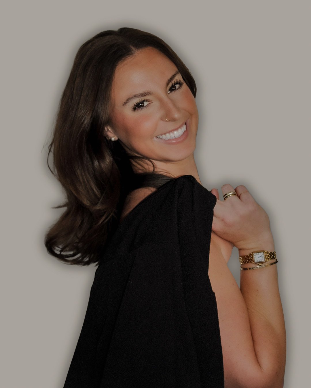 A woman with shoulder-length dark hair smiling, wearing a black top with the shoulder cut out, a gold wristwatch, and jewelry, posing against a plain background.
