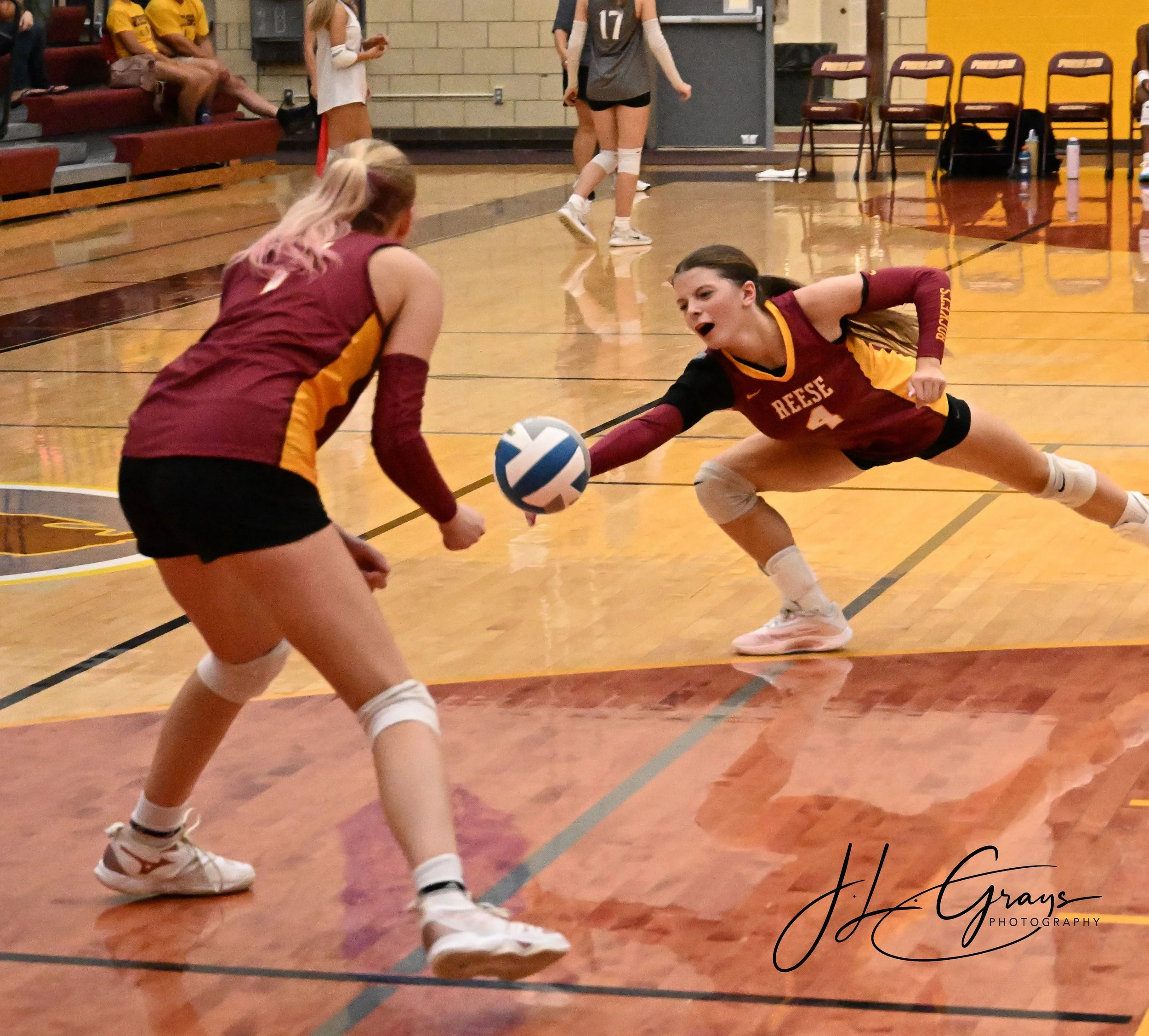 Two volleyball players on a gym floor, one with dark hair wearing maroon and yellow uniform with the name Reese, the other with blonde hair and pink highlights wearing similar uniform, both reaching for a volleyball.