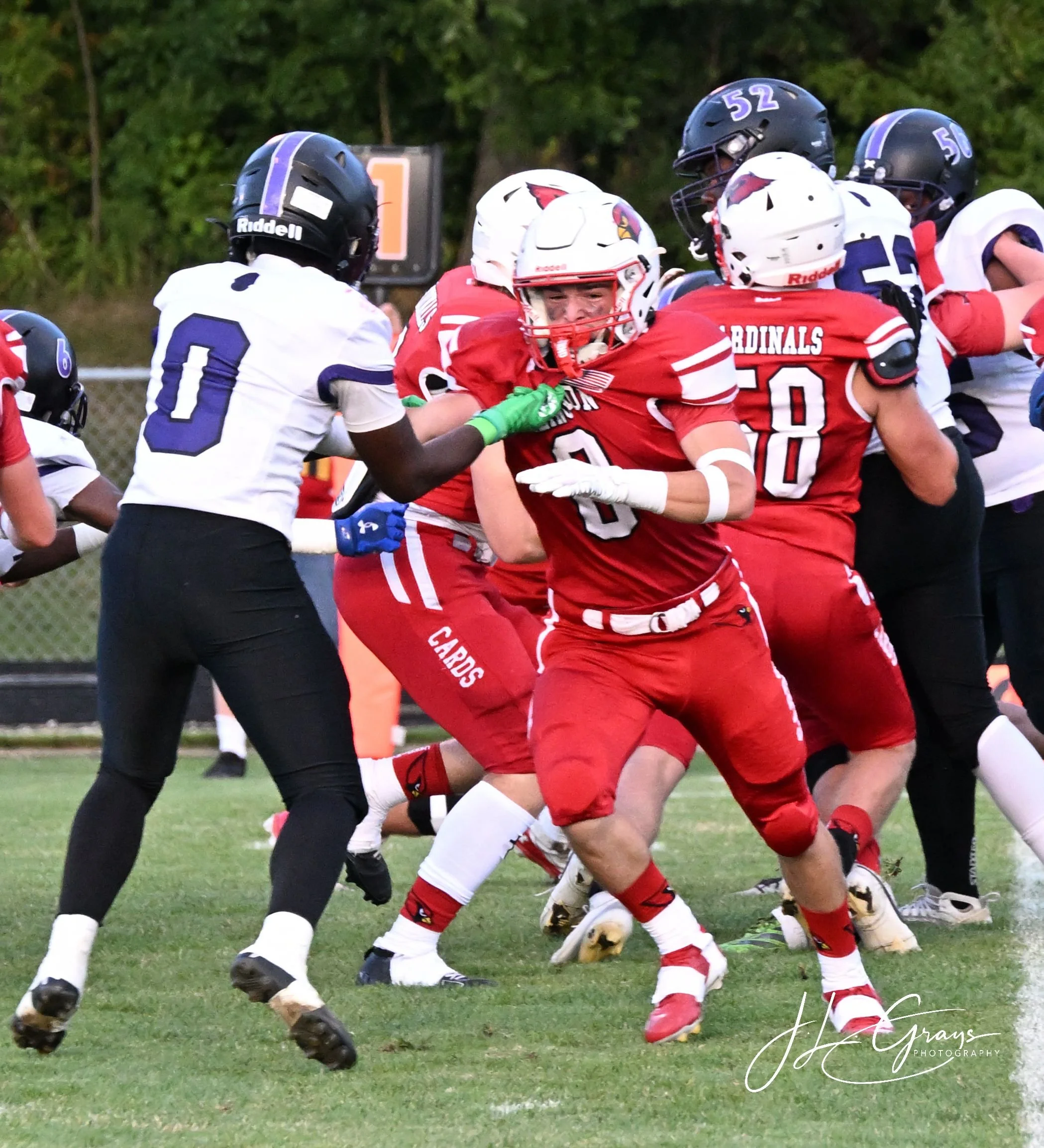 High school football game showing players in red and white uniforms and black helmets against players in white uniforms with blue and purple helmets, with players engaging in a tackle on a grassy field under a partly cloudy sky.