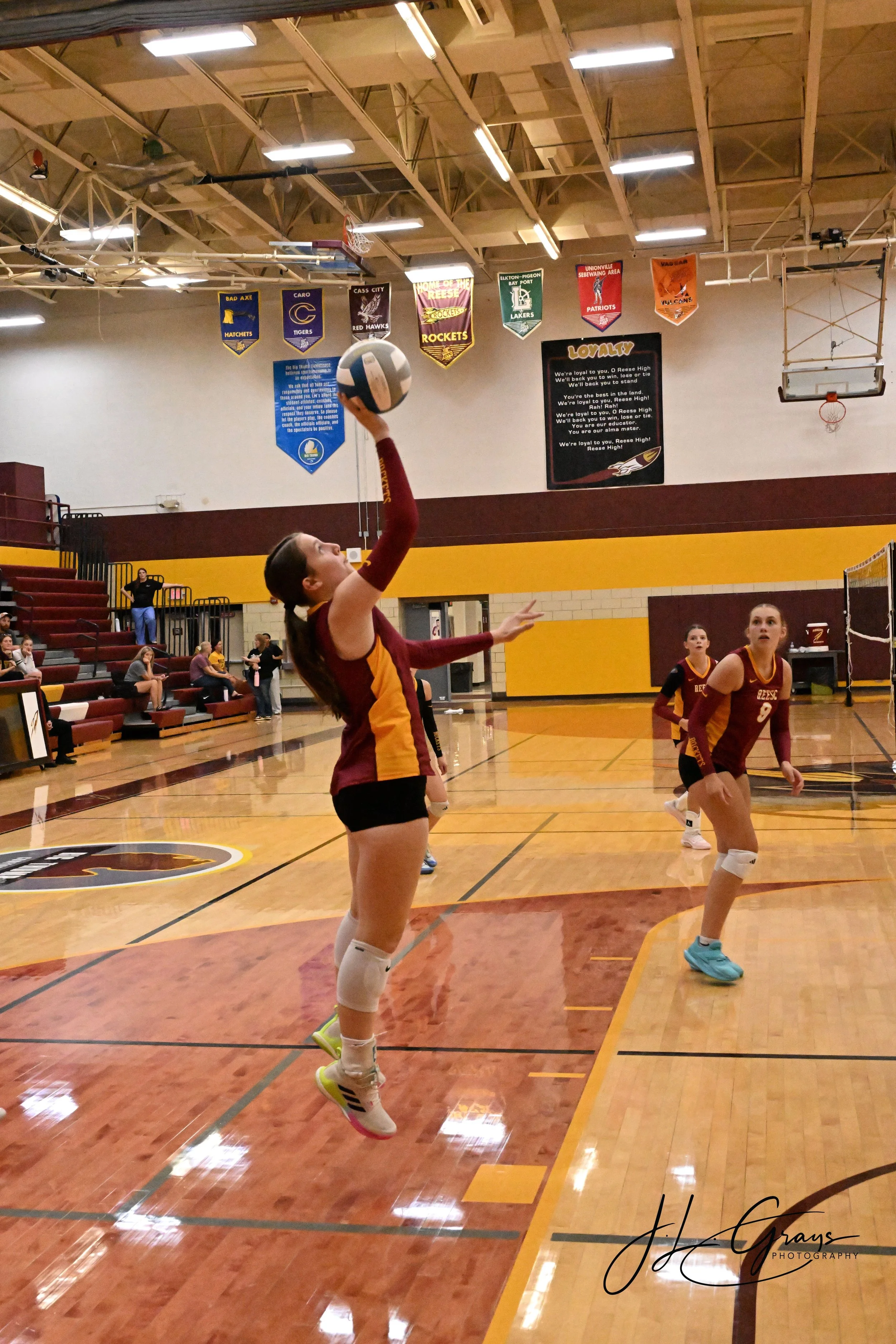 A girl in a maroon and gold volleyball uniform jumps in the gym, reaching out to hit a volleyball. She wears knee pads and colorful sneakers. Other players in similar uniforms are visible in the background, along with spectators seated on red and bla