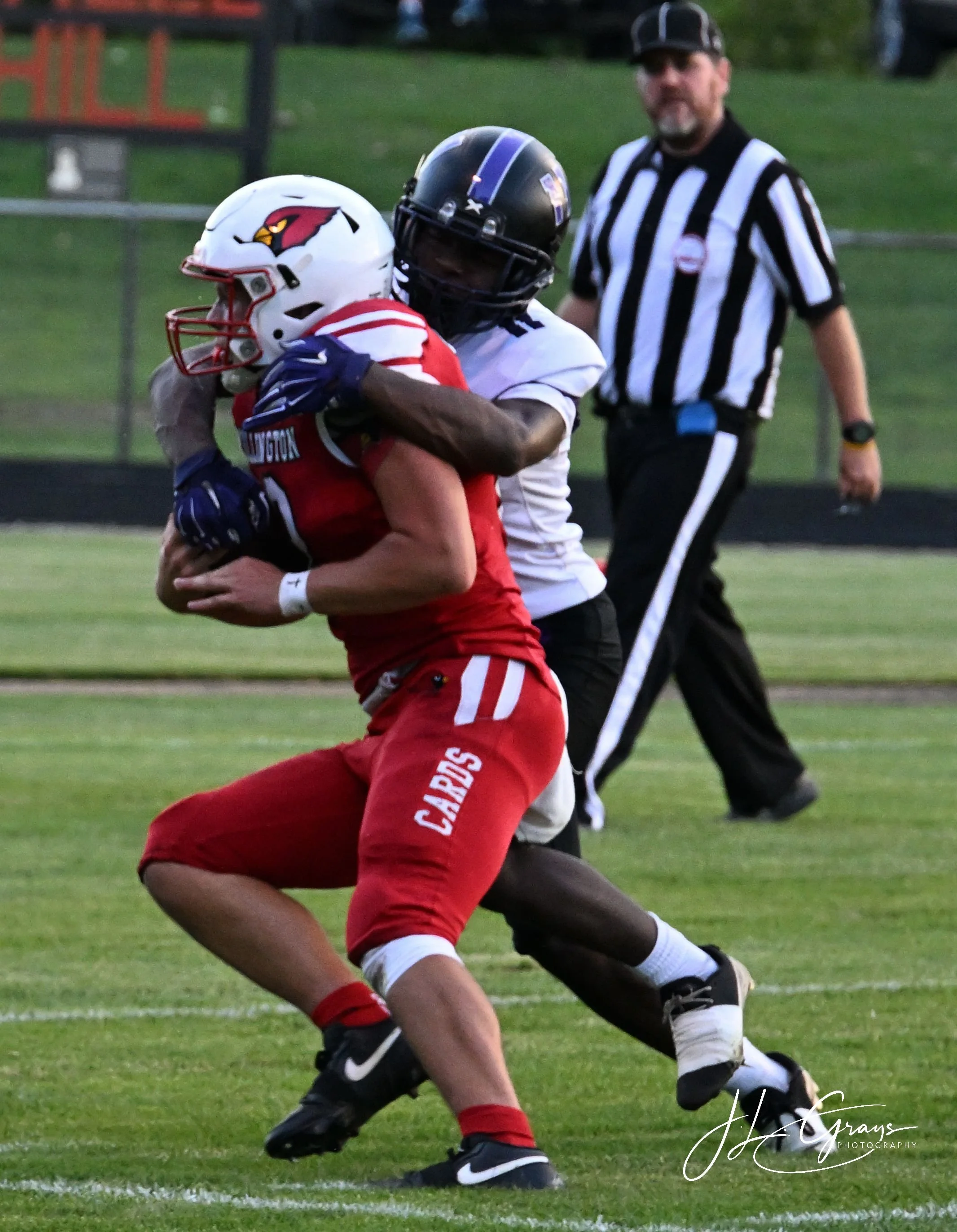 Two football players, one in a red uniform with a white helmet and the other in a white helmet, are engaged in a tackle on a football field. A referee stands in the background.