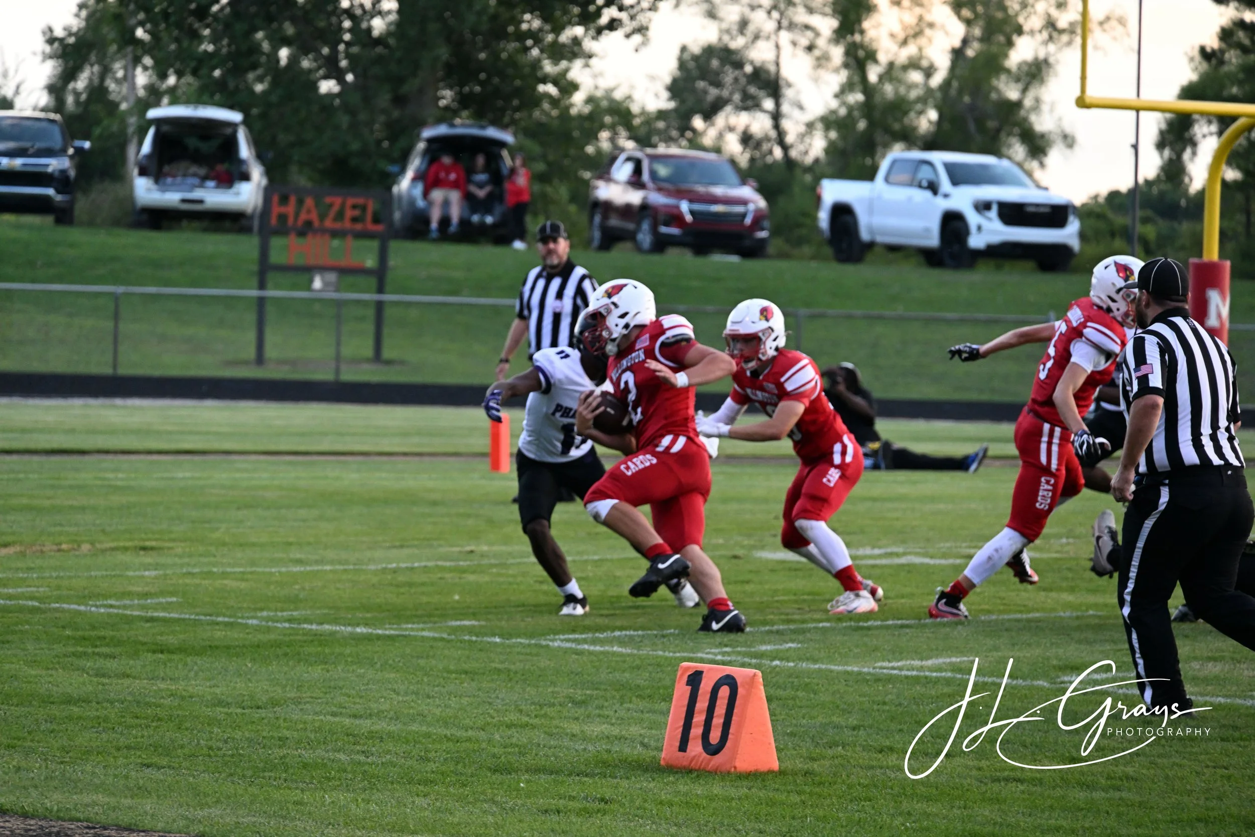 Football players in red and white uniforms playing on a football field during a game with a referee and a goalpost visible, spectators sitting in cars parked on a hill in the background.
